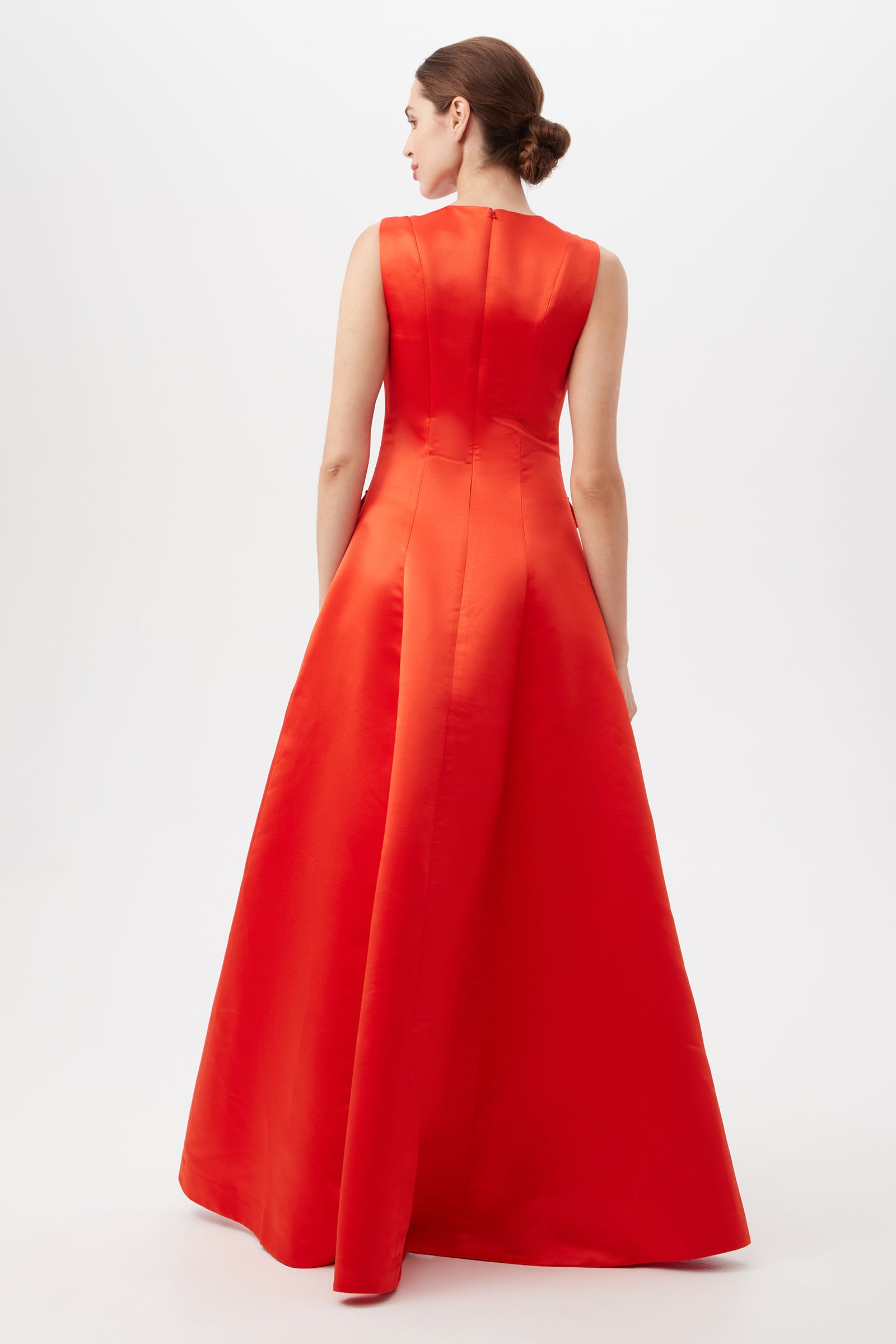 A woman with brown hair in a low bun models the Guinevere Dress, a sleeveless, floor-length bright red gown. She faces away from the camera, displaying the back of the dress against a plain white background.