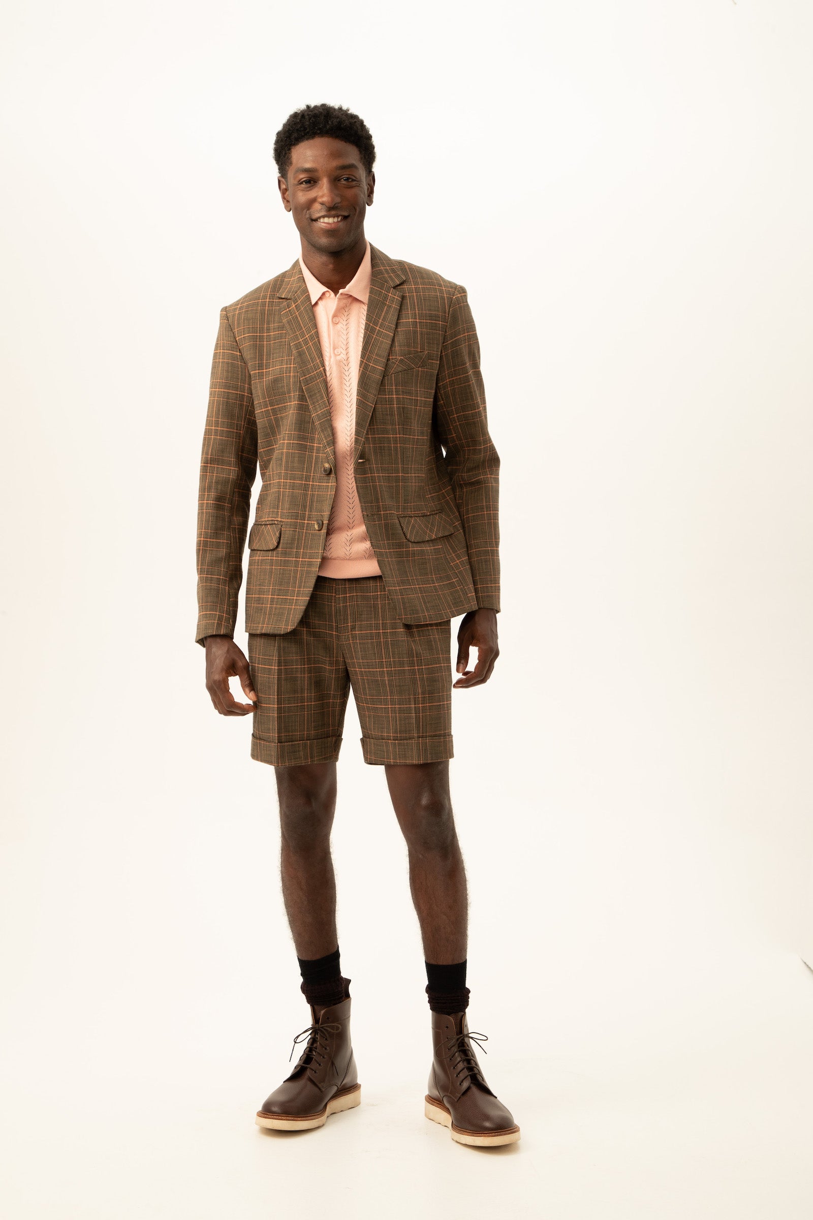 A man smiles wearing the Thurston 2 Blazer in Chaka Check Plaid with matching shorts, a light pink collared shirt, dark socks, and brown lace-up boots against a plain white background.