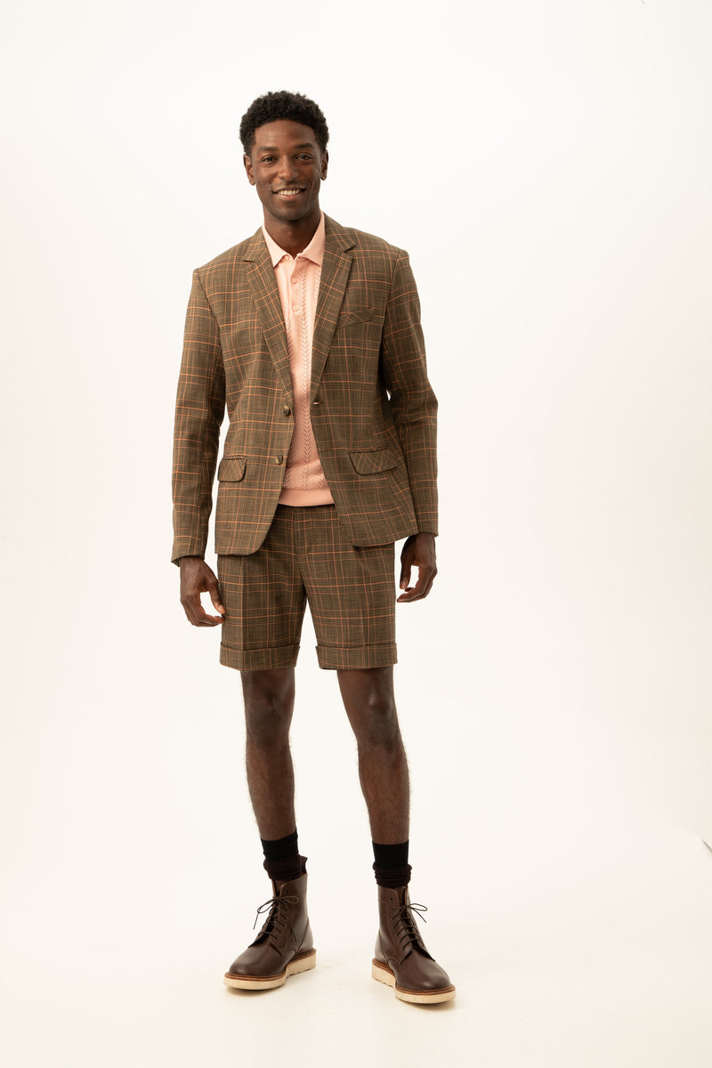 A man smiles wearing the Thurston 2 Blazer in Chaka Check Plaid with matching shorts, a light pink collared shirt, dark socks, and brown lace-up boots against a plain white background.