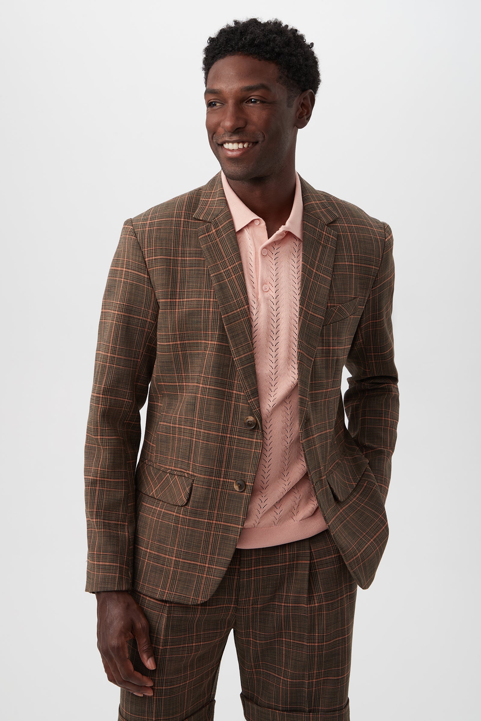 A man wears the Thurston 2 Blazer - Chaka Check Plaid (Multi) over a light pink textured polo shirt, standing with one hand in his pocket and smiling to the side against a plain white background.