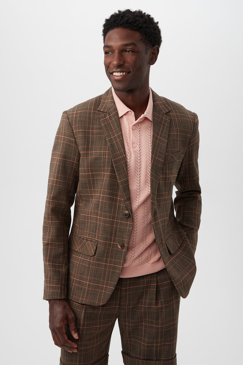 A man wears the Thurston 2 Blazer - Chaka Check Plaid (Multi) over a light pink textured polo shirt, standing with one hand in his pocket and smiling to the side against a plain white background.