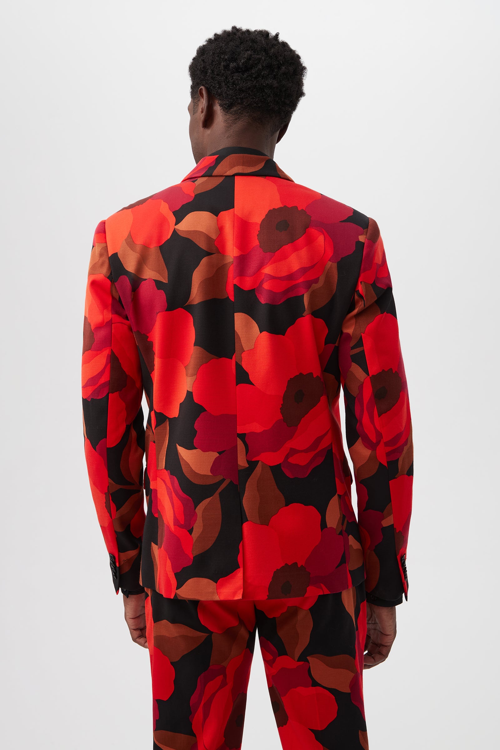 Someone is seen from behind wearing the Thurston Blazer - Fathala Faille, a matching suit jacket and pants set featuring a bold red, orange, and black floral pattern against a plain light background.