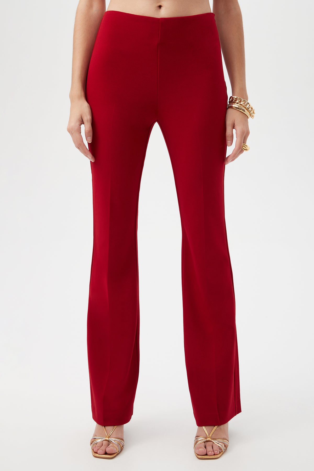 A person wearing Jacoba Crepe Knit Tailored Bootcut Pant in Roselle Red with gold strappy heels, gold bracelets, and rings, stands against a plain white background.