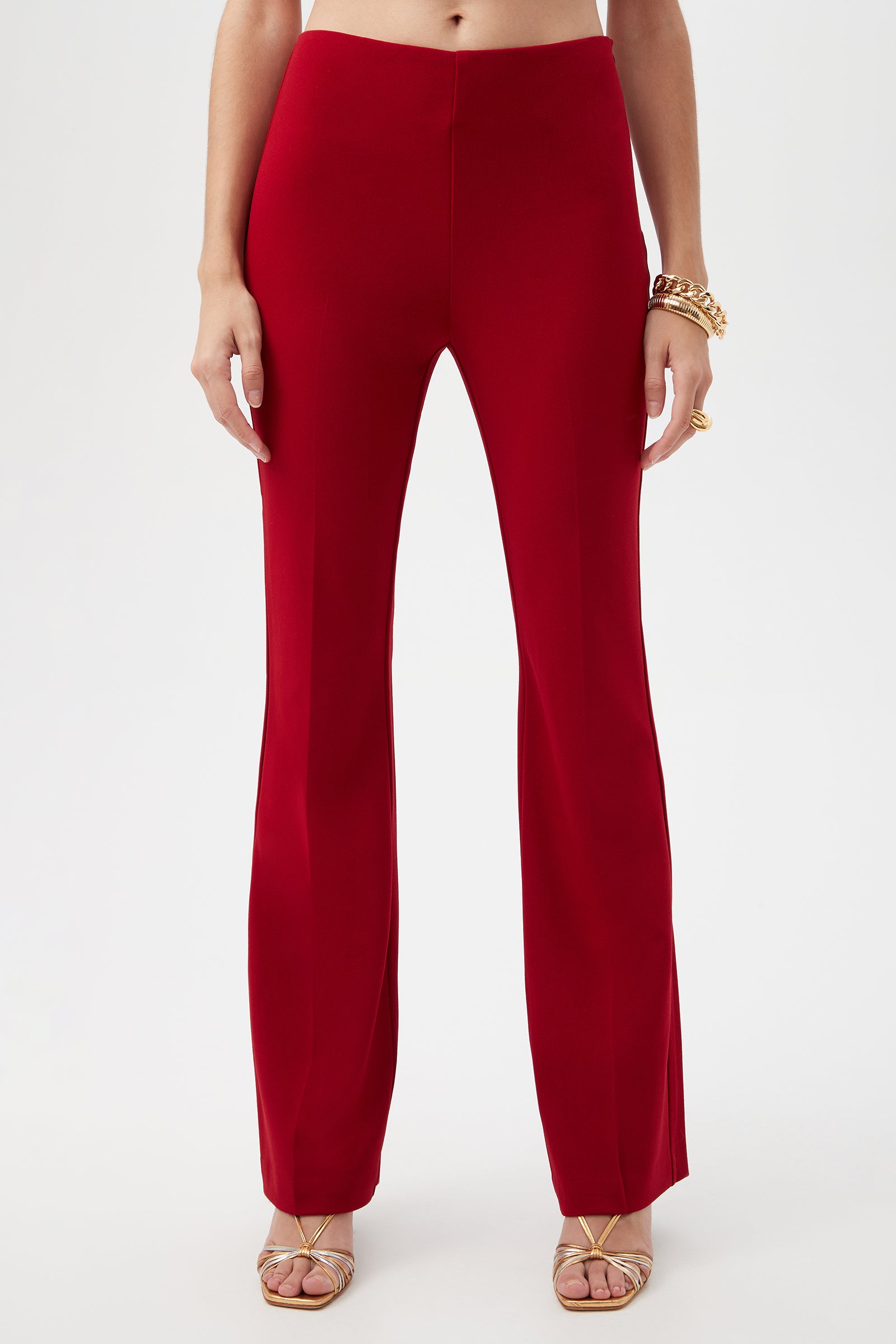 A person wearing Jacoba Crepe Knit Tailored Bootcut Pant in Roselle Red with gold strappy heels, gold bracelets, and rings, stands against a plain white background.