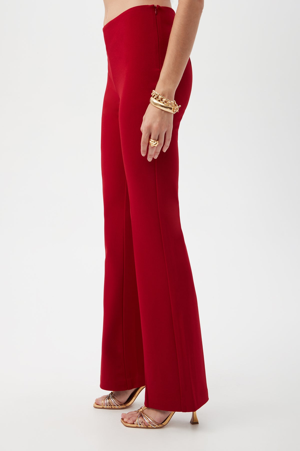 A person stands in profile against a plain white background, wearing the Jacoba Crepe Knit Tailored Bootcut Pant in bright red, paired with gold strappy heels and gold bracelets.
