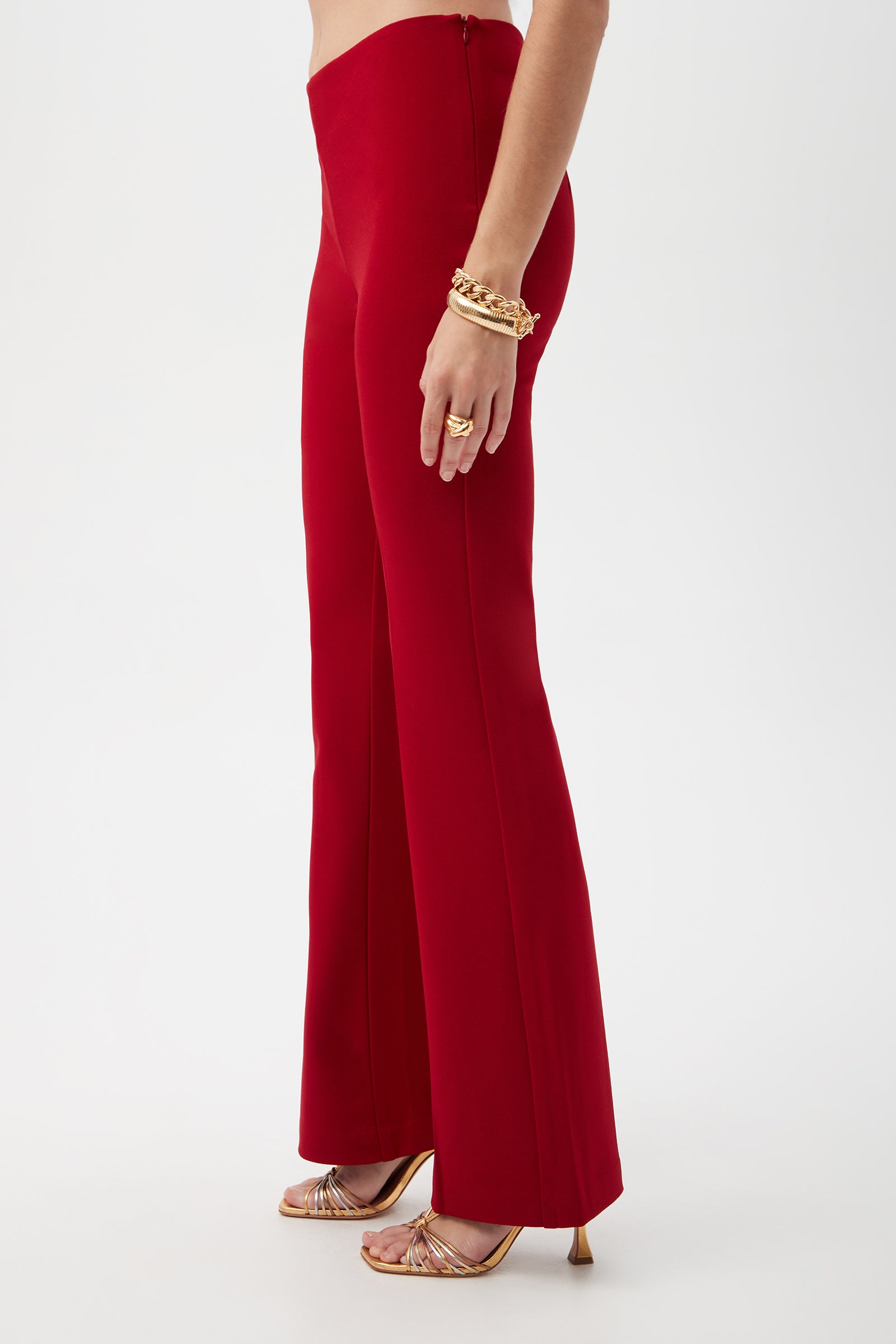 A person stands in profile against a plain white background, wearing the Jacoba Crepe Knit Tailored Bootcut Pant in bright red, paired with gold strappy heels and gold bracelets.