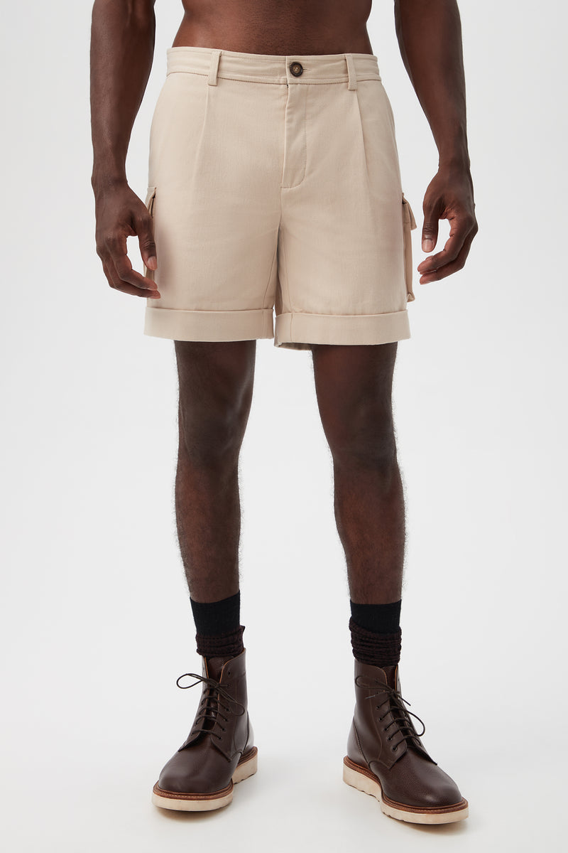 A person stands against a plain background wearing Westlake Cargo Short in Sahara Stone, featuring side pockets, paired with black socks and brown lace-up boots. The image is cropped from the chest to the feet.