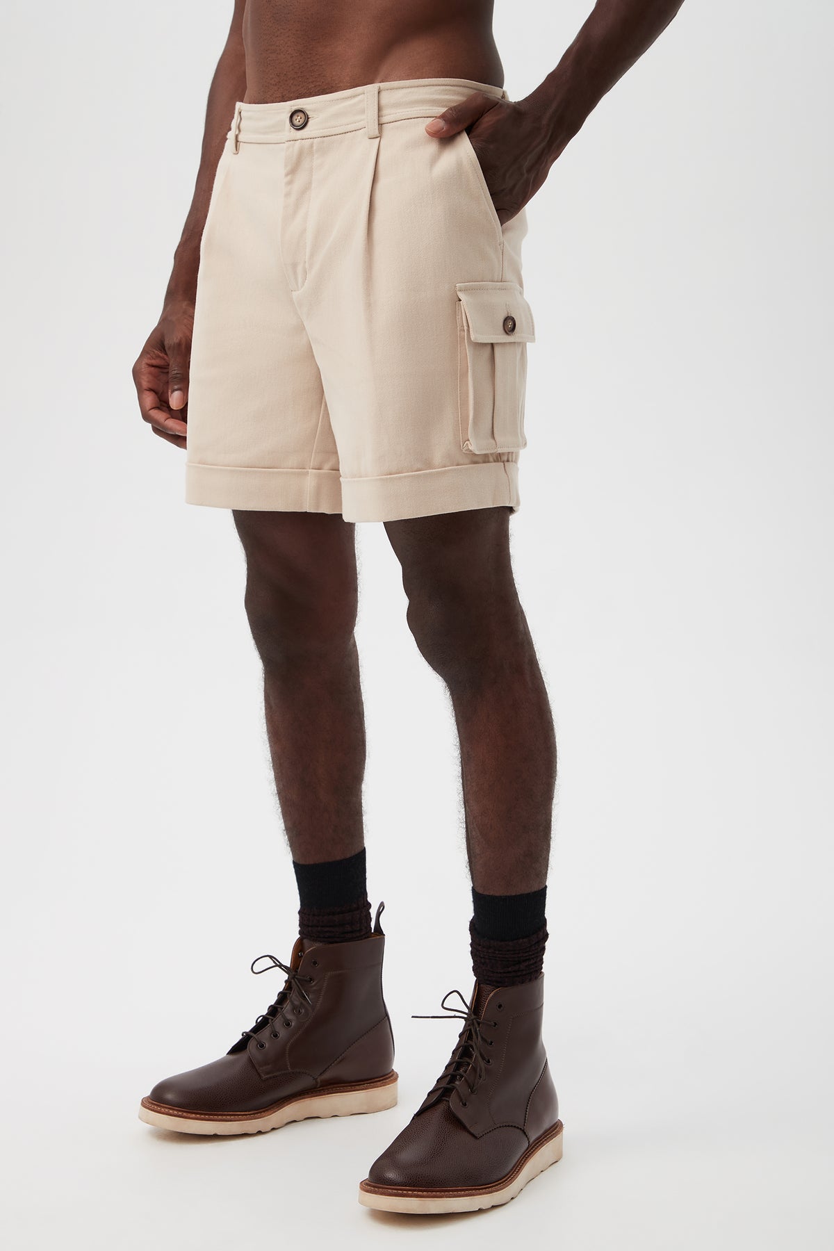 A person wearing Westlake Cargo Short - Brushed Cotton Twill in beige with a side pocket, paired with black socks and dark brown lace-up boots, standing against a plain white background.