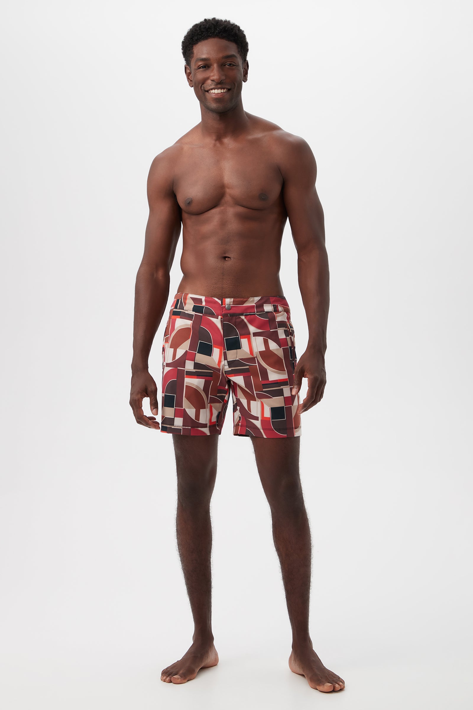 A smiling man stands barefoot against a plain white background, wearing the Honolulu Board Short - Galerie Arte featuring a colorful geometric pattern and no shirt.