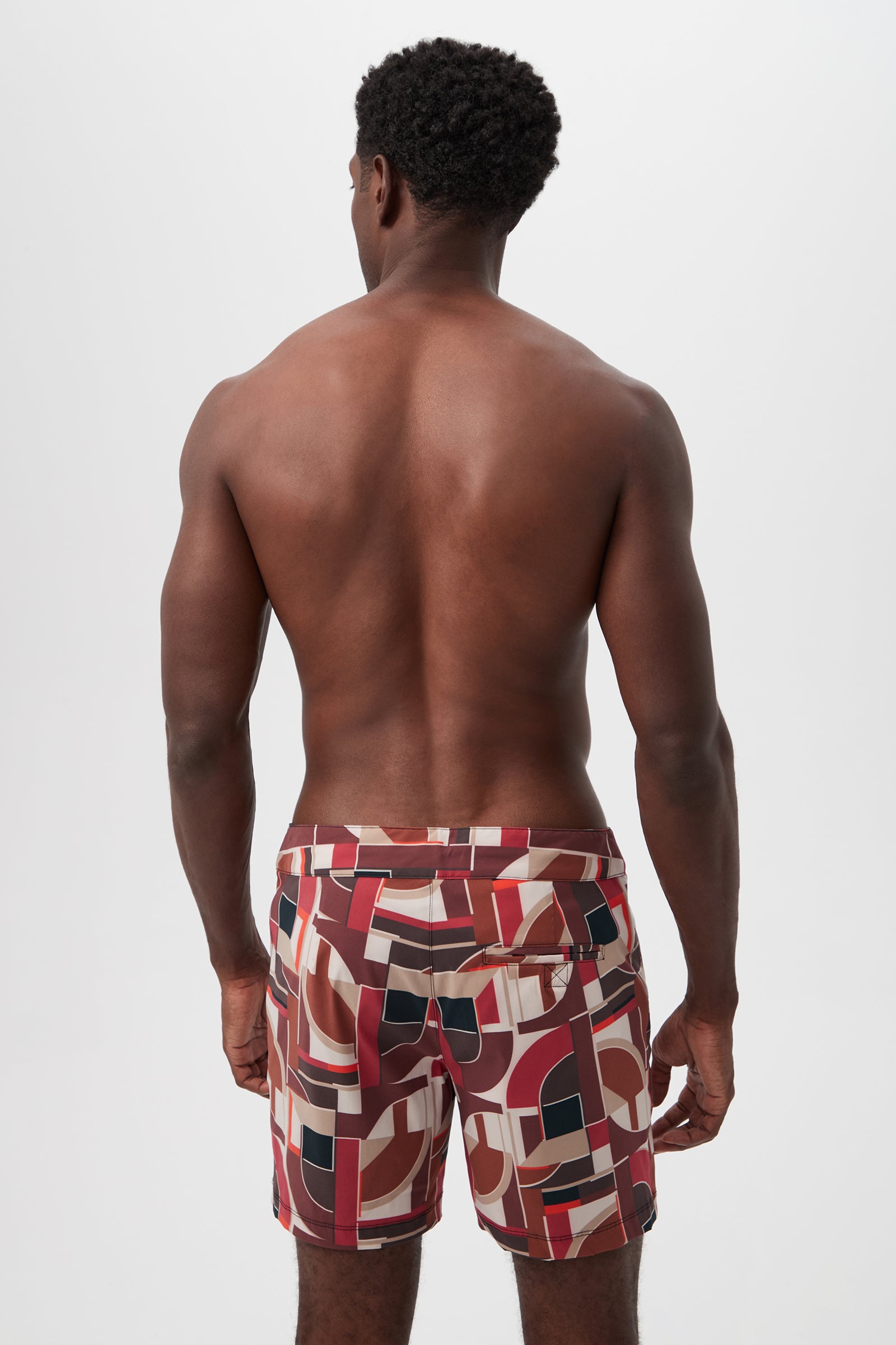 A shirtless man faces away, wearing the Honolulu Board Short - Galerie Arte featuring red, beige, black, and white geometric patterns against a plain white background.