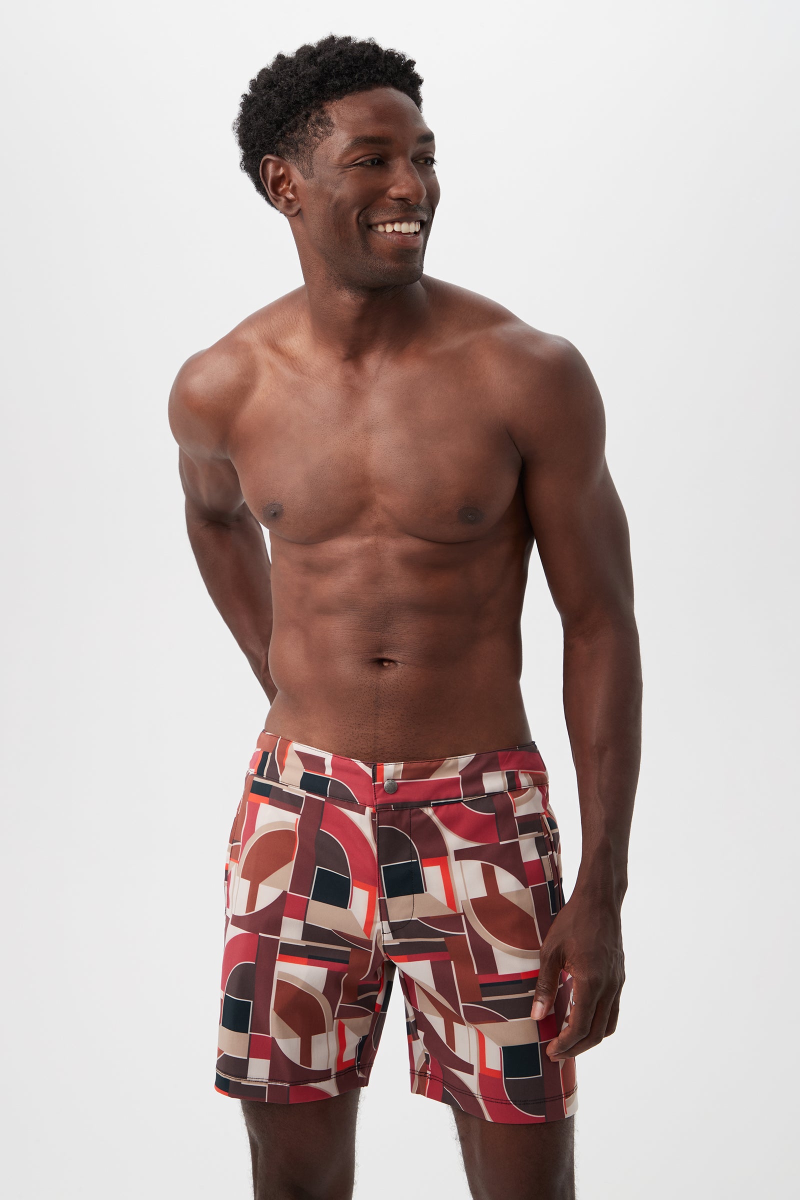 A smiling, shirtless man stands against a white background wearing the Honolulu Board Short - Galerie Arte, featuring colorful geometric patterns in red, brown, black, and white.