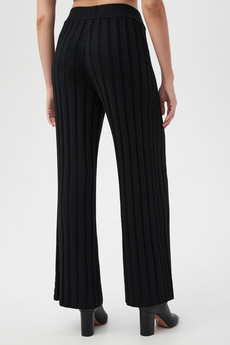 A person stands facing away, wearing the Pearce 2 Pant—high-waisted, wide-leg black ribbed knit pants—with black heeled ankle boots against a plain white background.