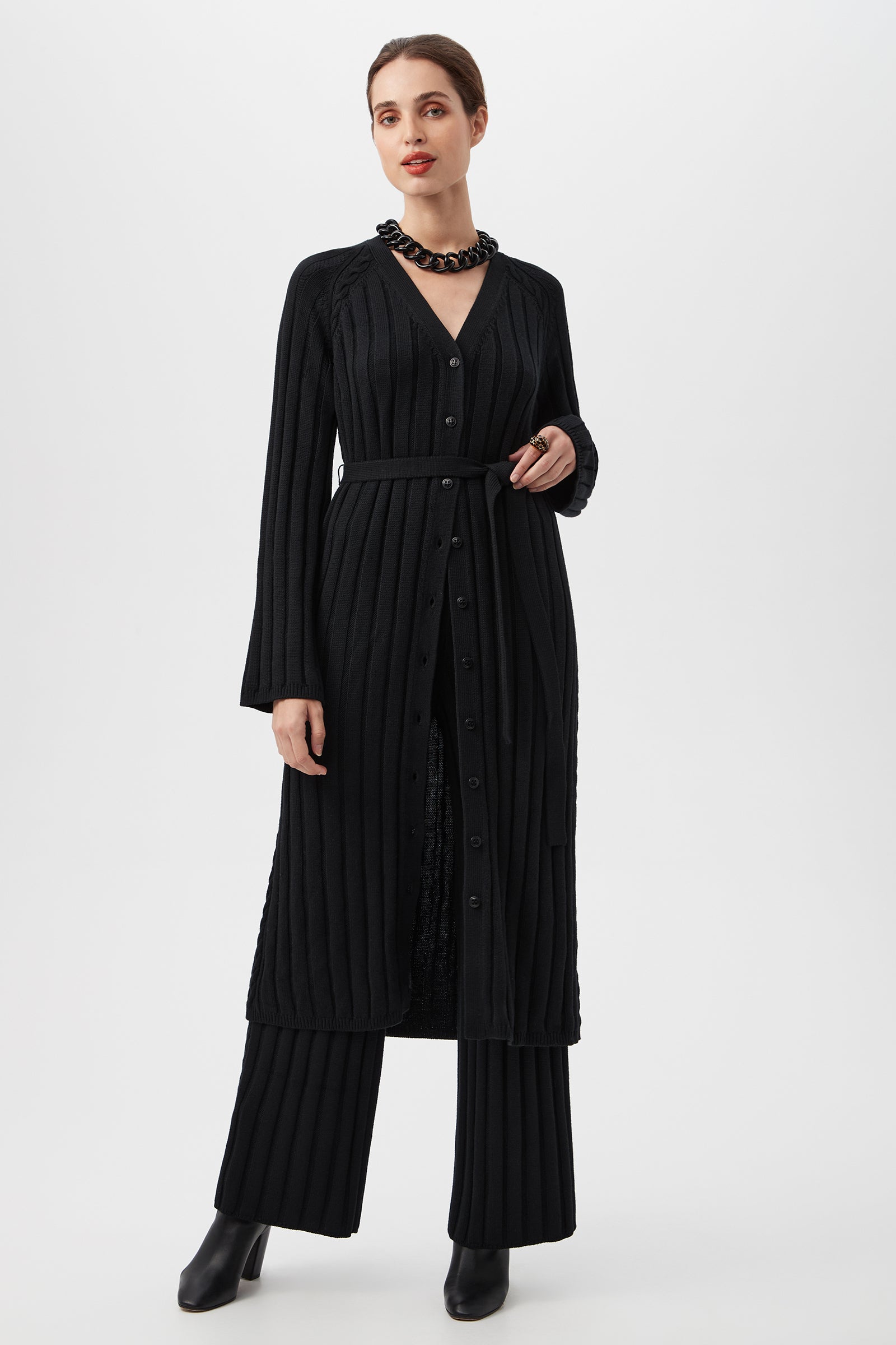 A woman stands against a white background wearing the black St. Helena Cardigan, styled with matching black wide-leg pants, black boots, and a chunky black necklace.