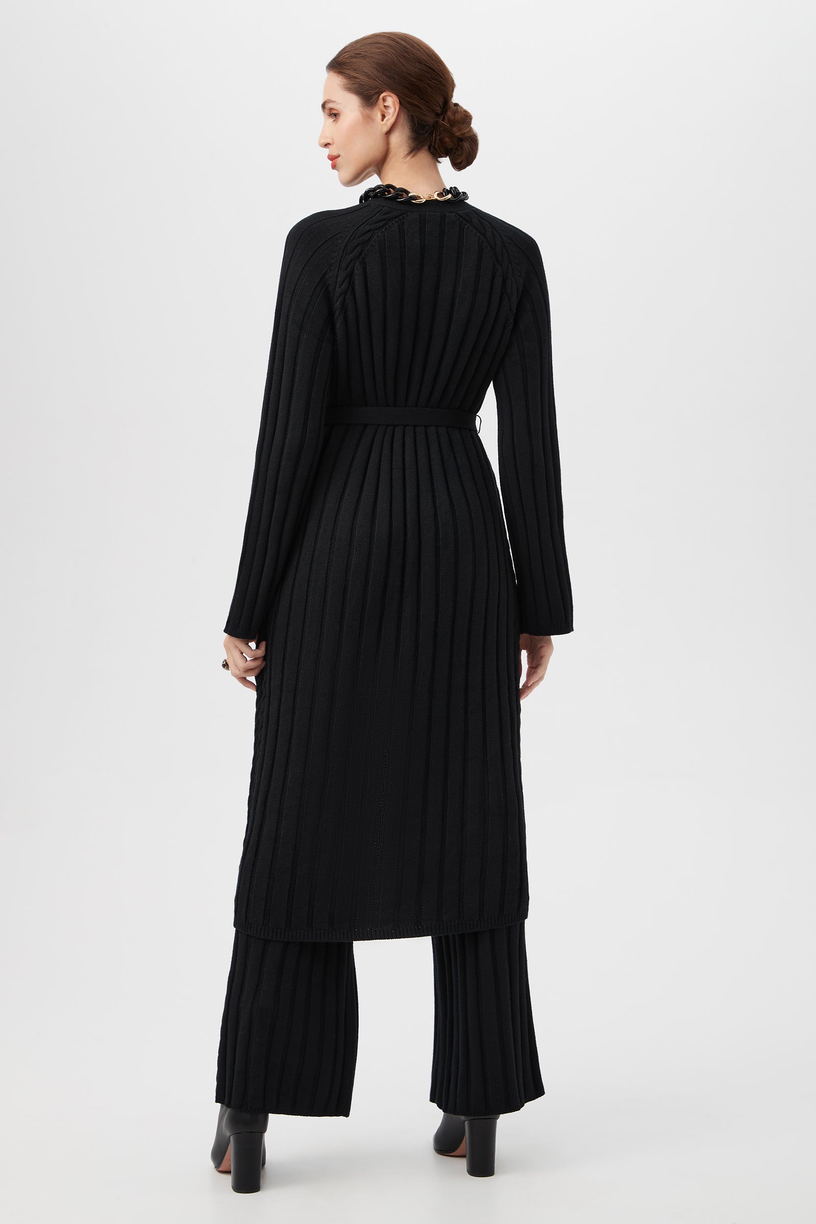 A woman stands facing away, wearing the long black St. Helena Cardigan with matching wide-leg pants and black heeled boots. Her hair is styled in a neat low bun against a plain white background.