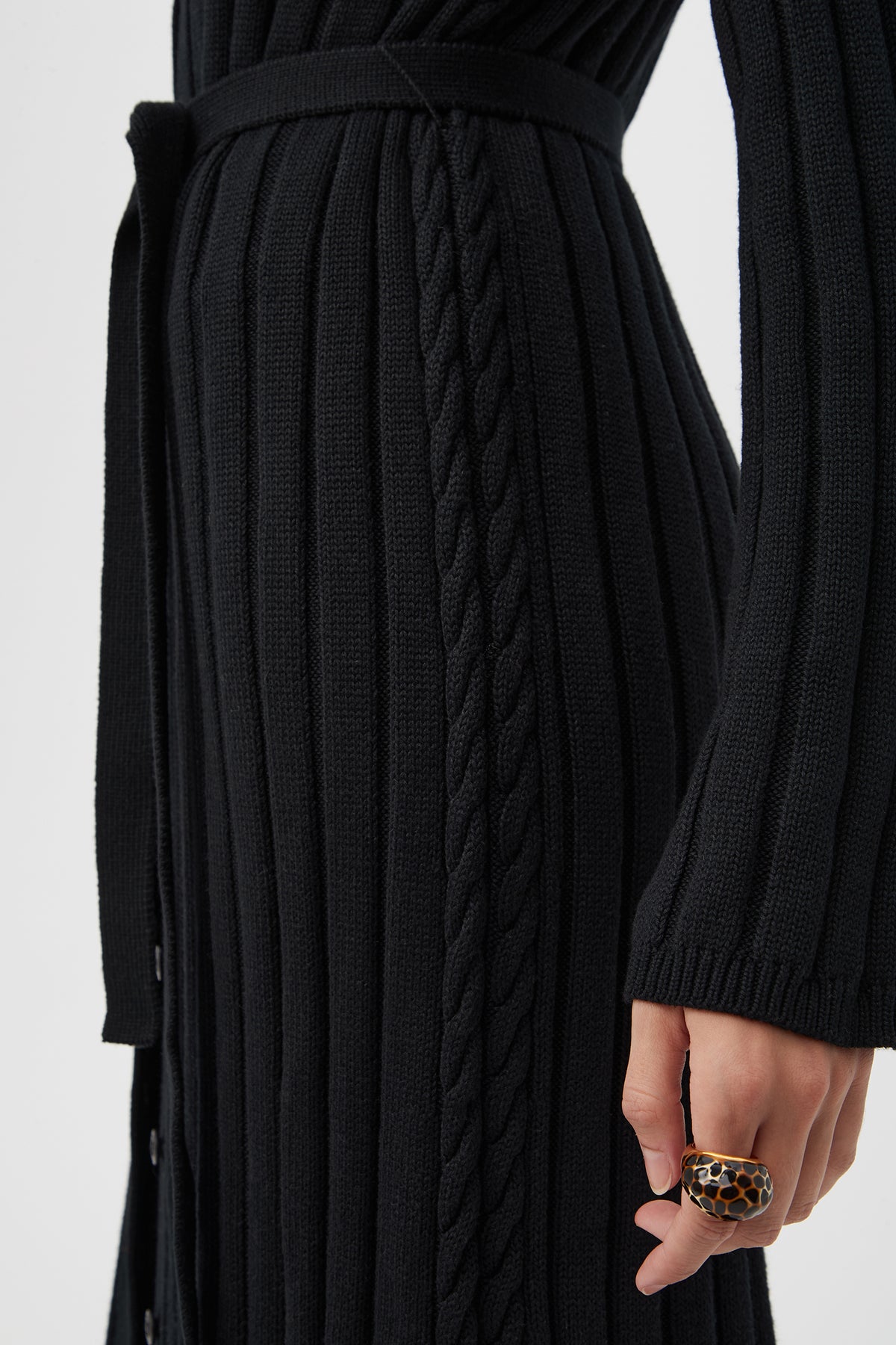 A close-up of a person wearing the St. Helena Cardigan, featuring cable knit details and a belted waist, with part of their arm and hand visible, accessorized with a chunky ring.