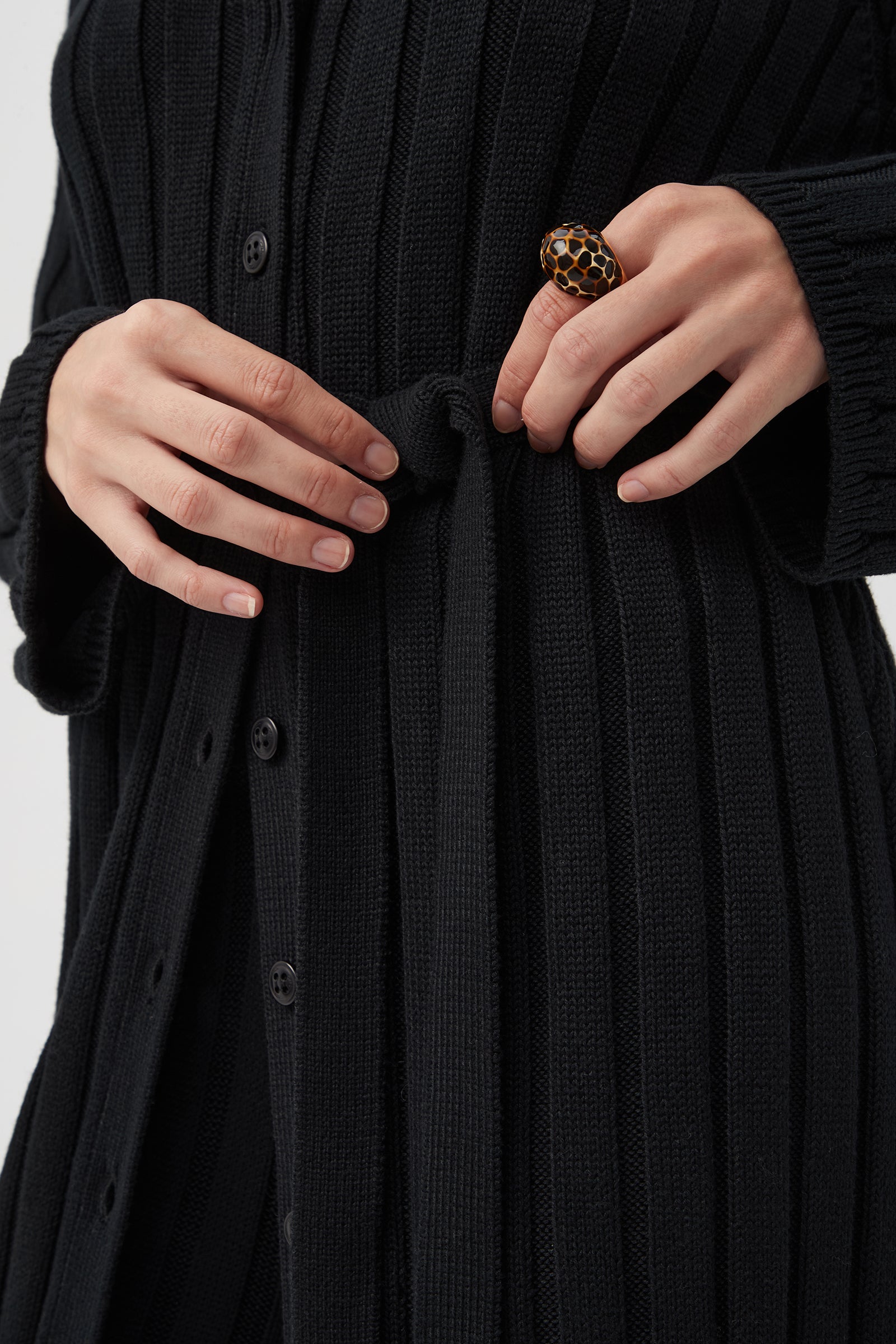 A person ties the belt of a St. Helena Cardigan, a black ribbed button-down style, while wearing a large dark-and-gold patterned ring on their right hand. The photo is cropped to show only the torso and hands.