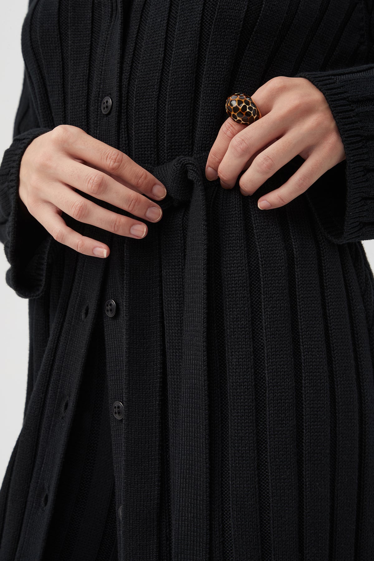 A person ties the belt of a St. Helena Cardigan, a black ribbed button-down style, while wearing a large dark-and-gold patterned ring on their right hand. The photo is cropped to show only the torso and hands.