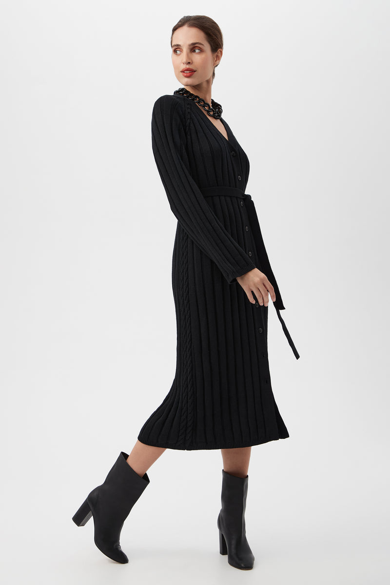 A woman stands turned slightly to the side against a plain white background, wearing the St. Helena Cardigan—a long-sleeved, black, ribbed midi dress with front buttons and a waist tie—paired with black heeled ankle boots.