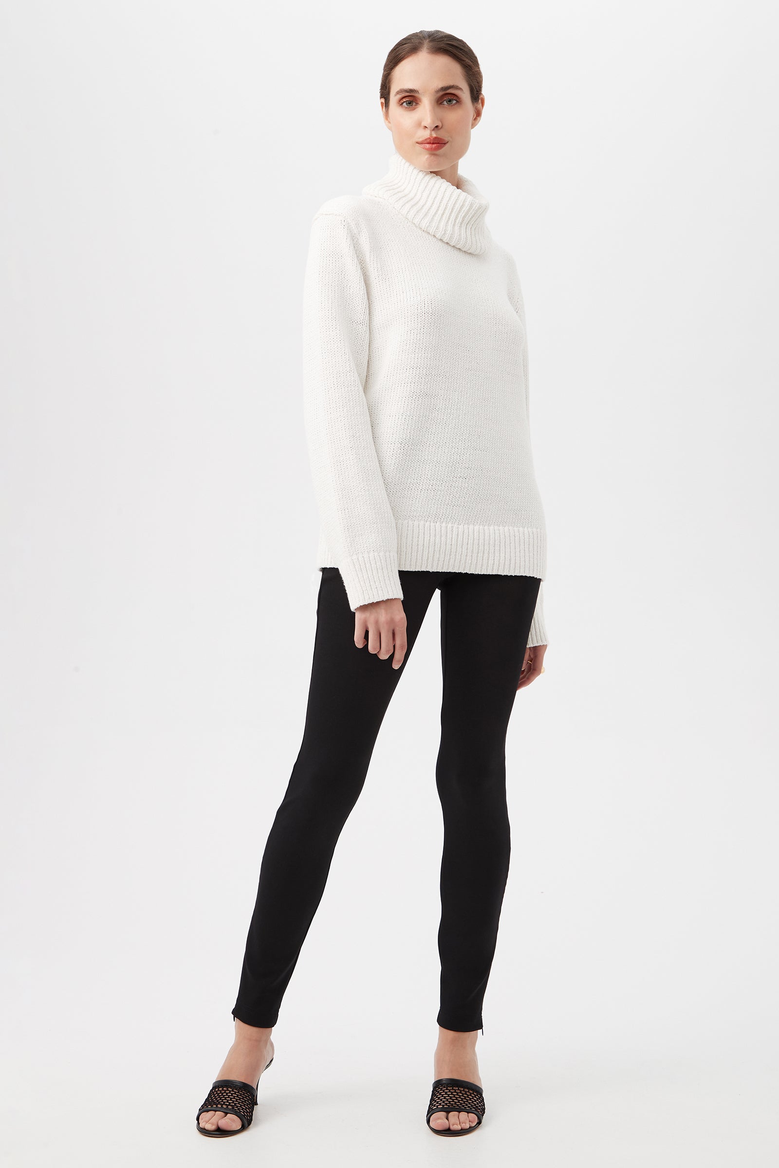 A person stands against a plain white background, wearing the Kalahari Sweater, black leggings, and black open-toe sandals. Their hair is pulled back and they are looking straight ahead.