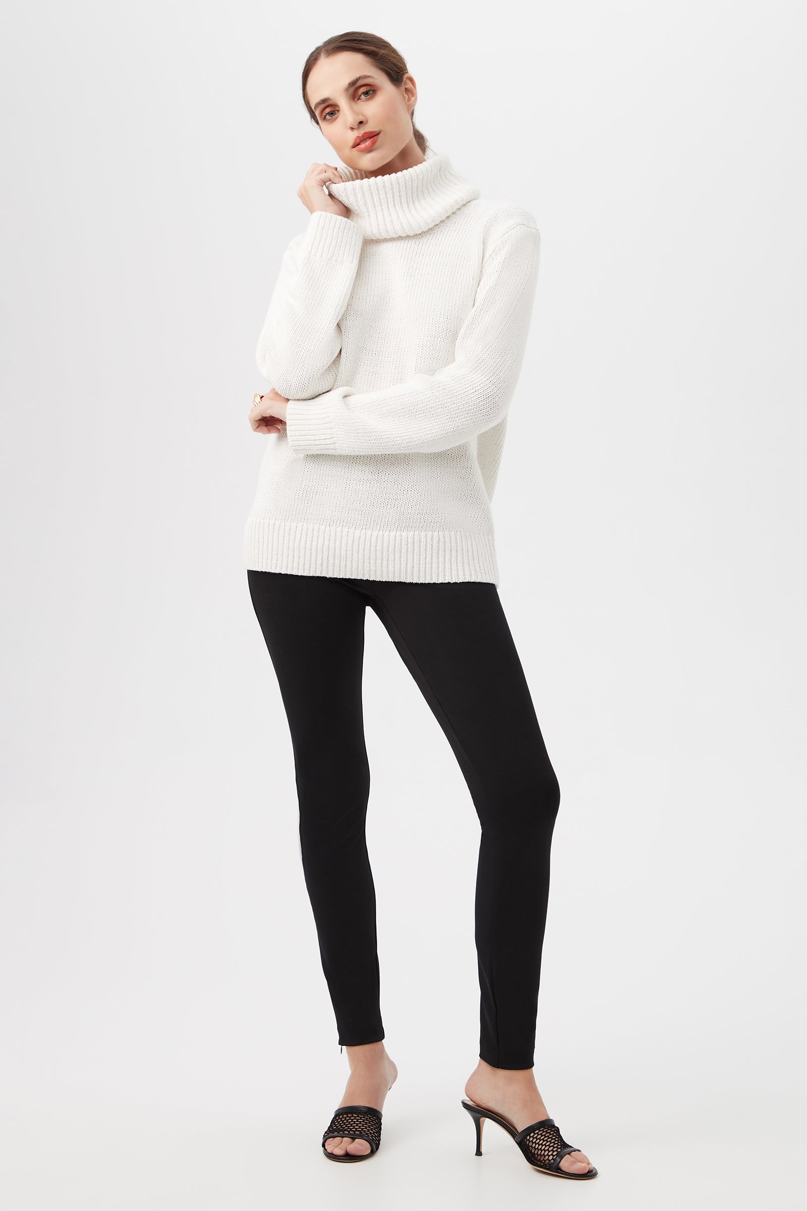 A woman poses confidently against a plain background wearing the Kalahari Sweater, a chunky white turtleneck, paired with black leggings and black open-toe heeled sandals. She rests her hand near her chin as she looks at the camera.