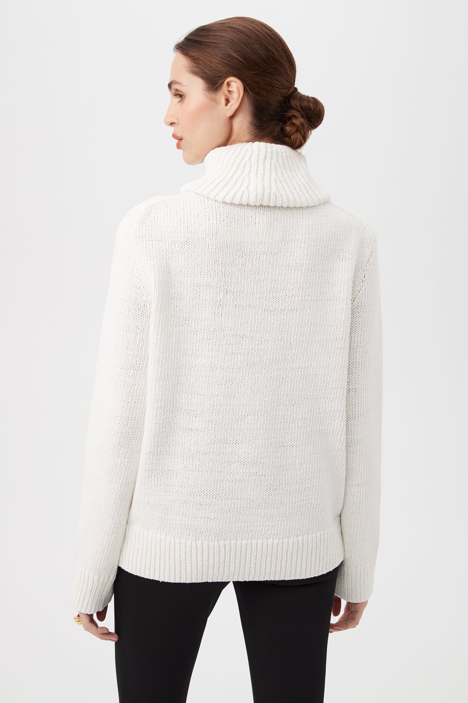 Woman with brown hair in a low bun wears the Kalahari Sweater, a white chunky knit turtleneck, paired with black pants, standing with her back to the camera against a plain light background.