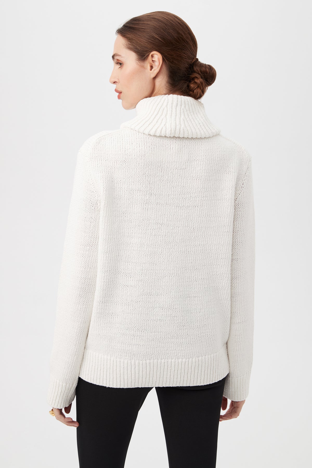 Woman with brown hair in a low bun wears the Kalahari Sweater, a white chunky knit turtleneck, paired with black pants, standing with her back to the camera against a plain light background.