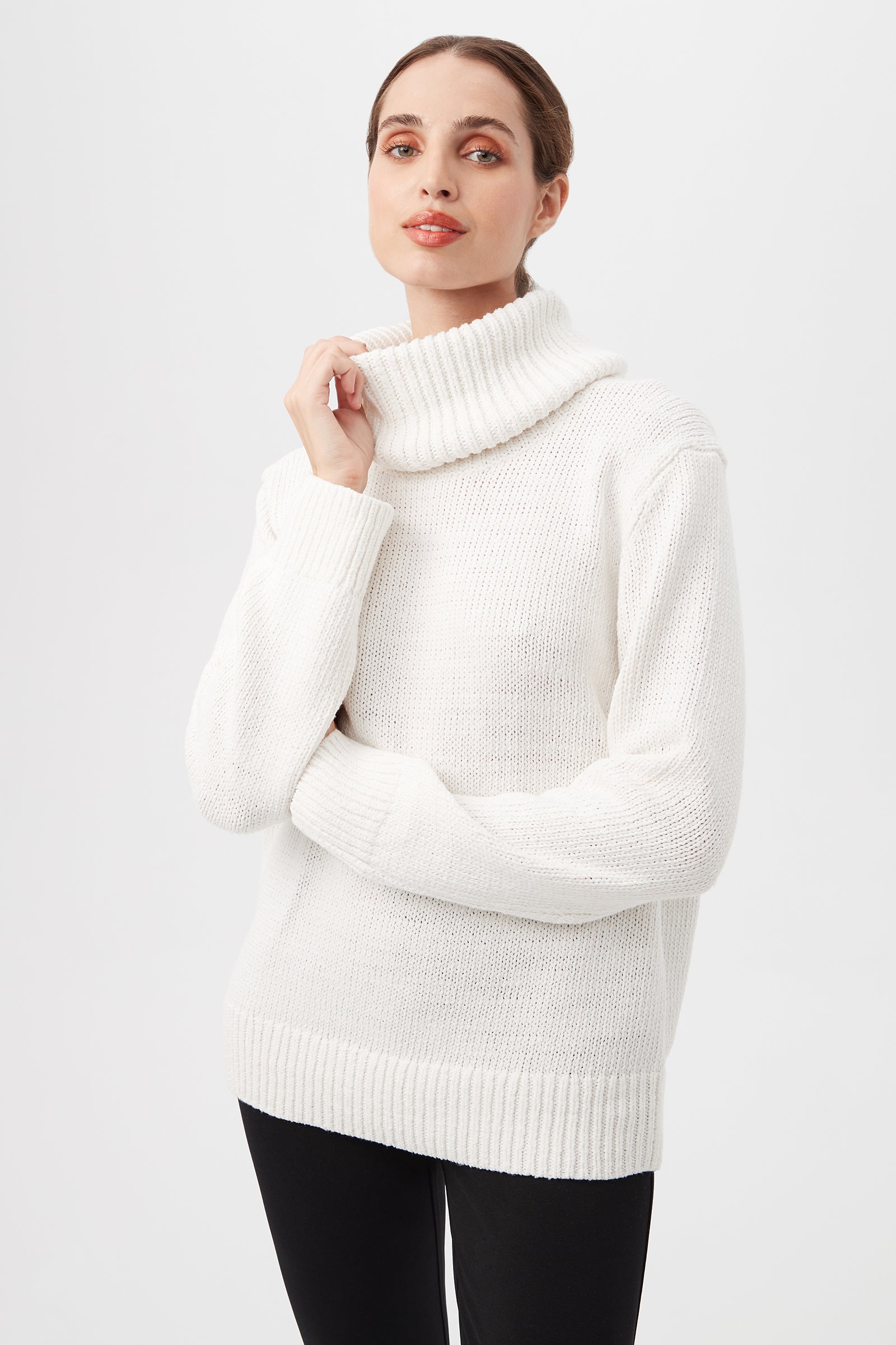 A woman wears the Kalahari Sweater in Winter White with black pants, standing against a plain light background, looking at the camera with a slight smile and one hand touching her collar.