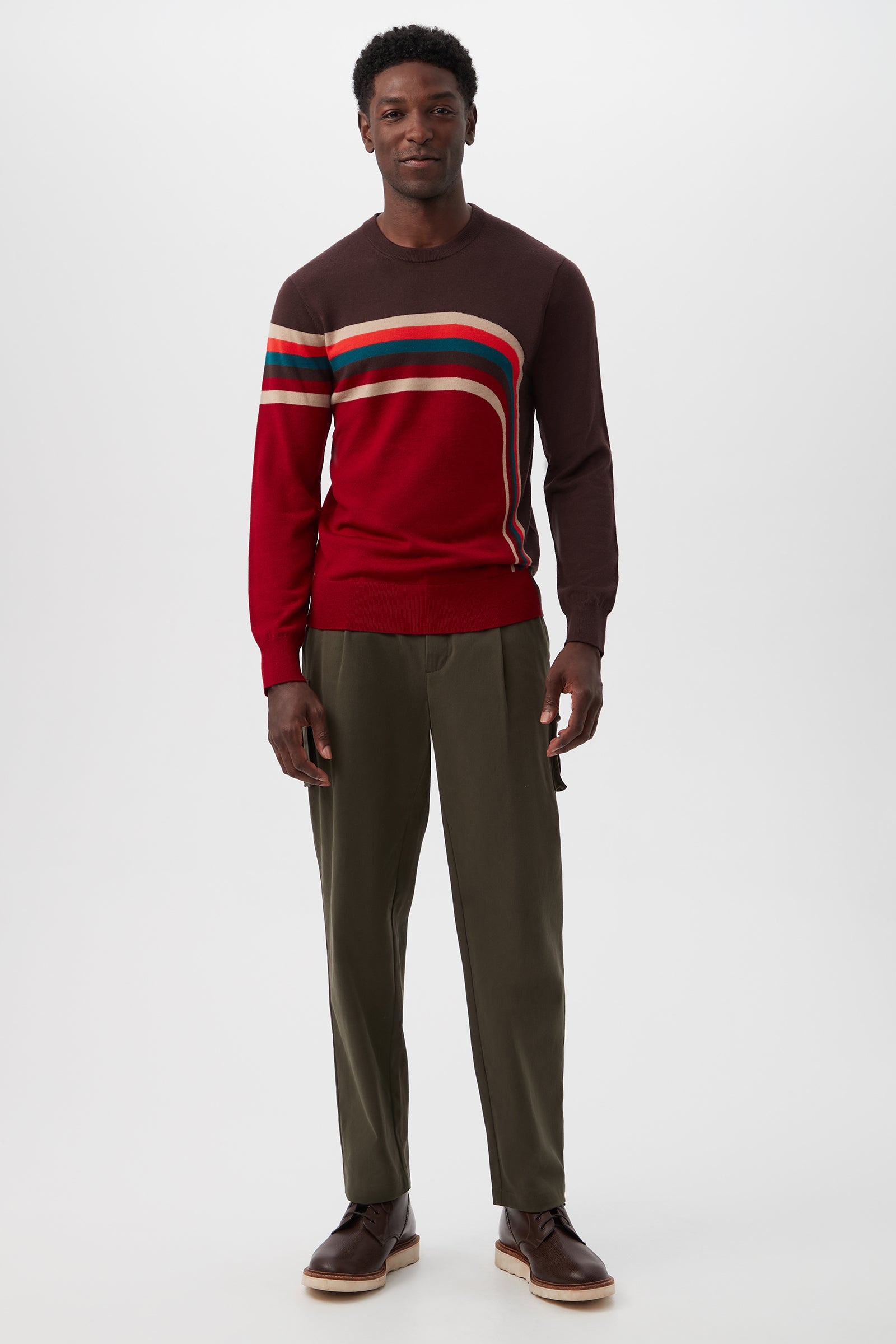 A man faces forward with a neutral expression, wearing the Rowan Crewneck Sweater in red and brown horizontal stripes, paired with olive green pants and brown shoes against a plain background.