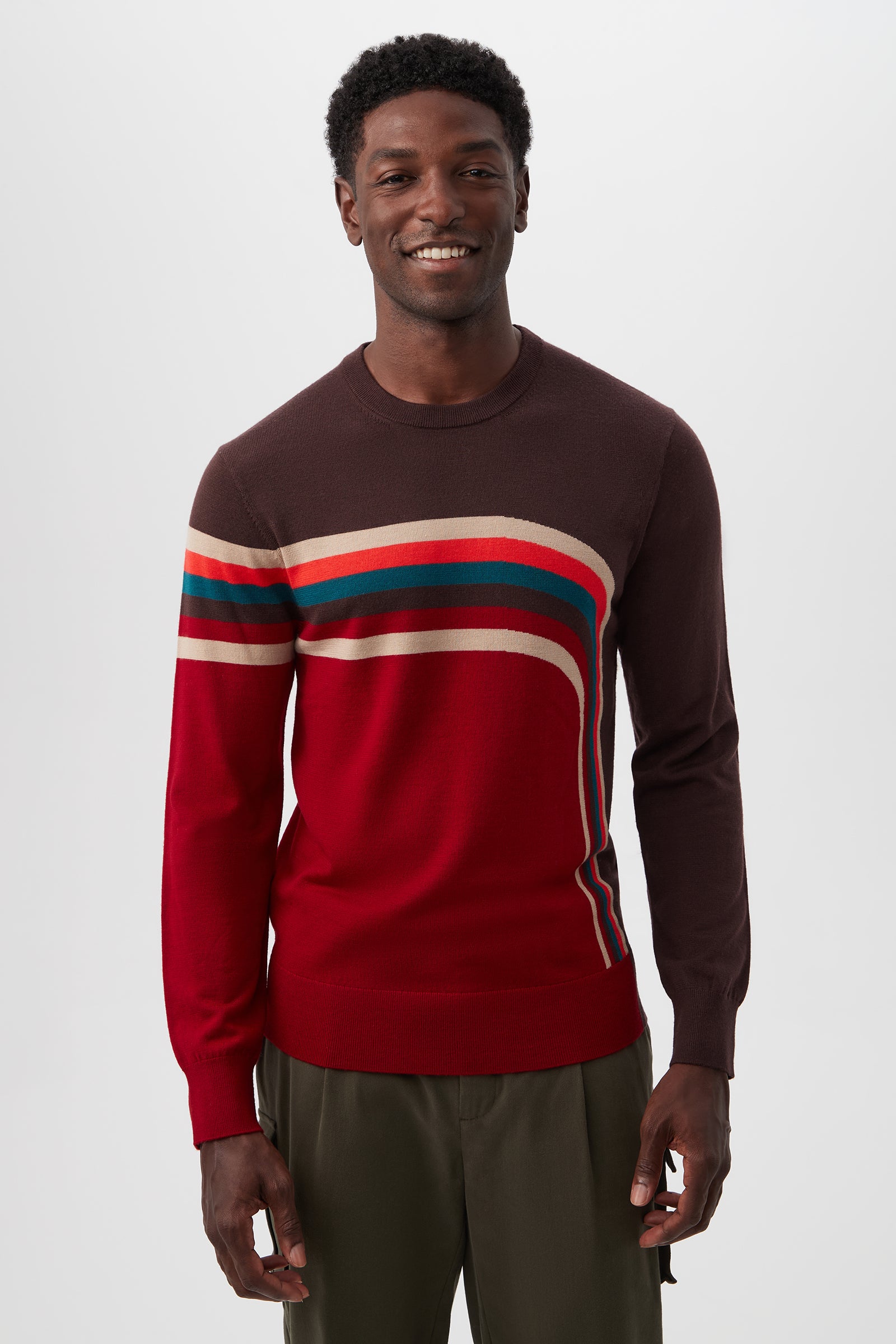 A man with short curly hair smiles in front of a plain white background, wearing the Rowan Crewneck Sweater - Merino Wool in Multi, featuring red, brown, beige, and dark green curved stripes, paired with olive green pants.