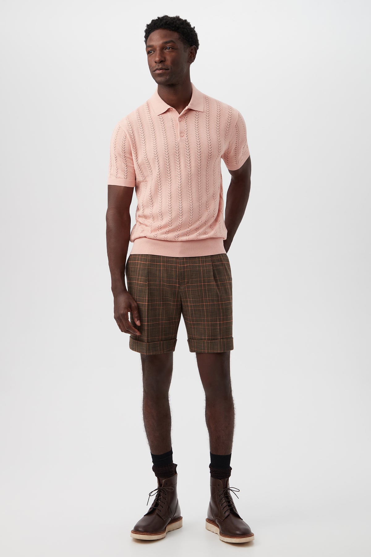 A man stands against a white background wearing brown Oliver 2 Short - Chaka Check Plaid shorts, a light pink short-sleeve polo, black socks, and brown lace-up boots. His posture is relaxed with hands by his sides.