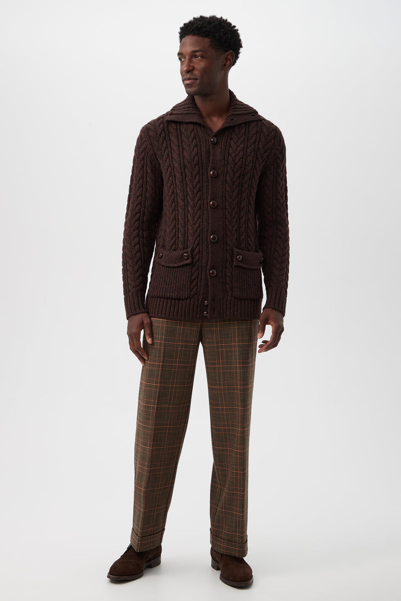 A man stands against a plain background wearing the Raleigh Sweater Coat - Merino Wool, featuring a dark brown cable-knit design with buttons and front pockets, paired with plaid brown trousers and dark brown shoes. He looks slightly to the side, relaxed.
