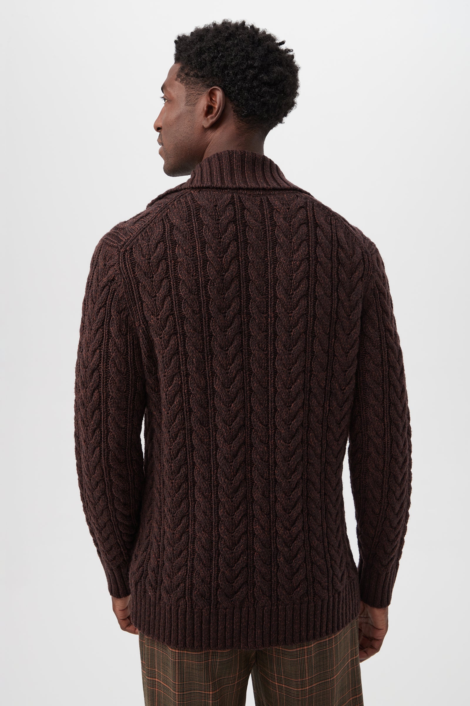 A man with short curly hair faces away, wearing the Raleigh Sweater Coat in brown merino wool featuring a chunky cable-knit and shawl collar, paired with brown plaid trousers against a plain light background.