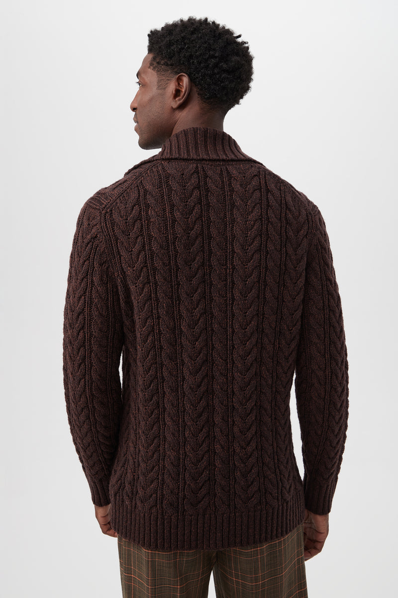 A man with short curly hair faces away, wearing the Raleigh Sweater Coat in brown merino wool featuring a chunky cable-knit and shawl collar, paired with brown plaid trousers against a plain light background.