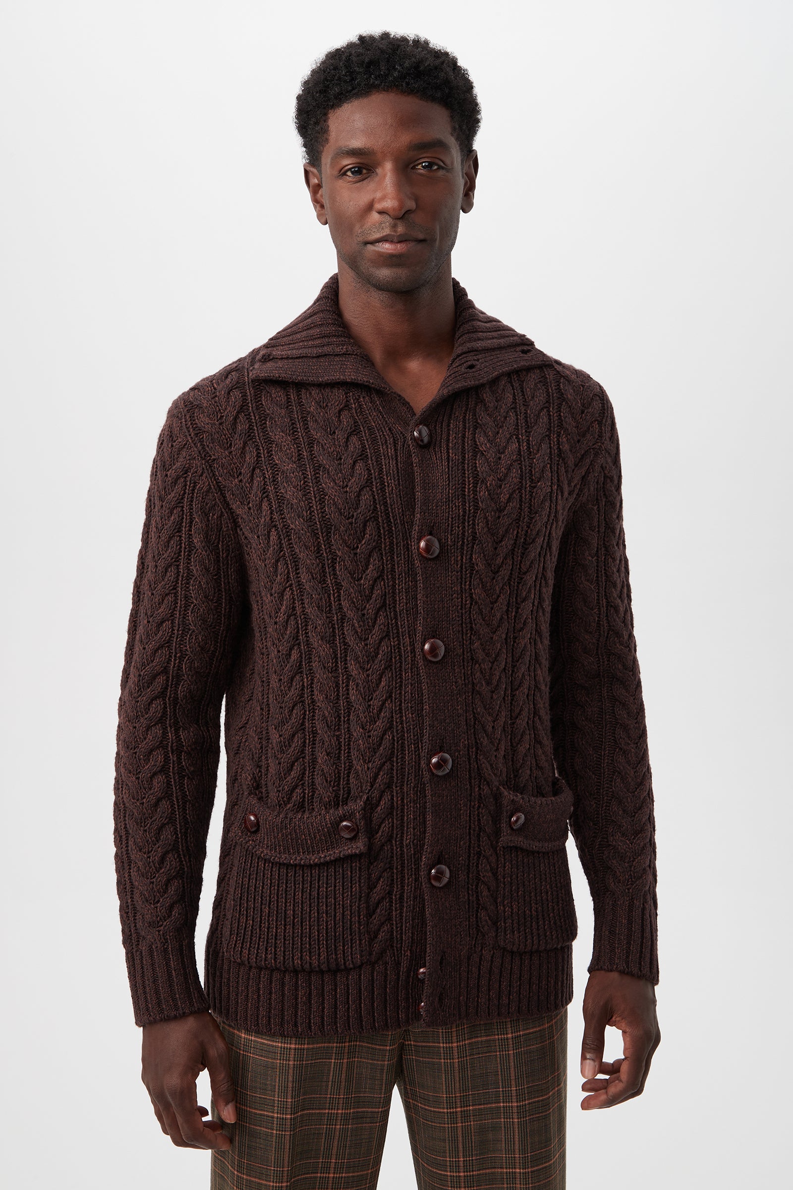 A man wearing the Raleigh Sweater Coat in dark brown merino wool with front pockets and buttons, paired with plaid pants, stands against a plain white background.