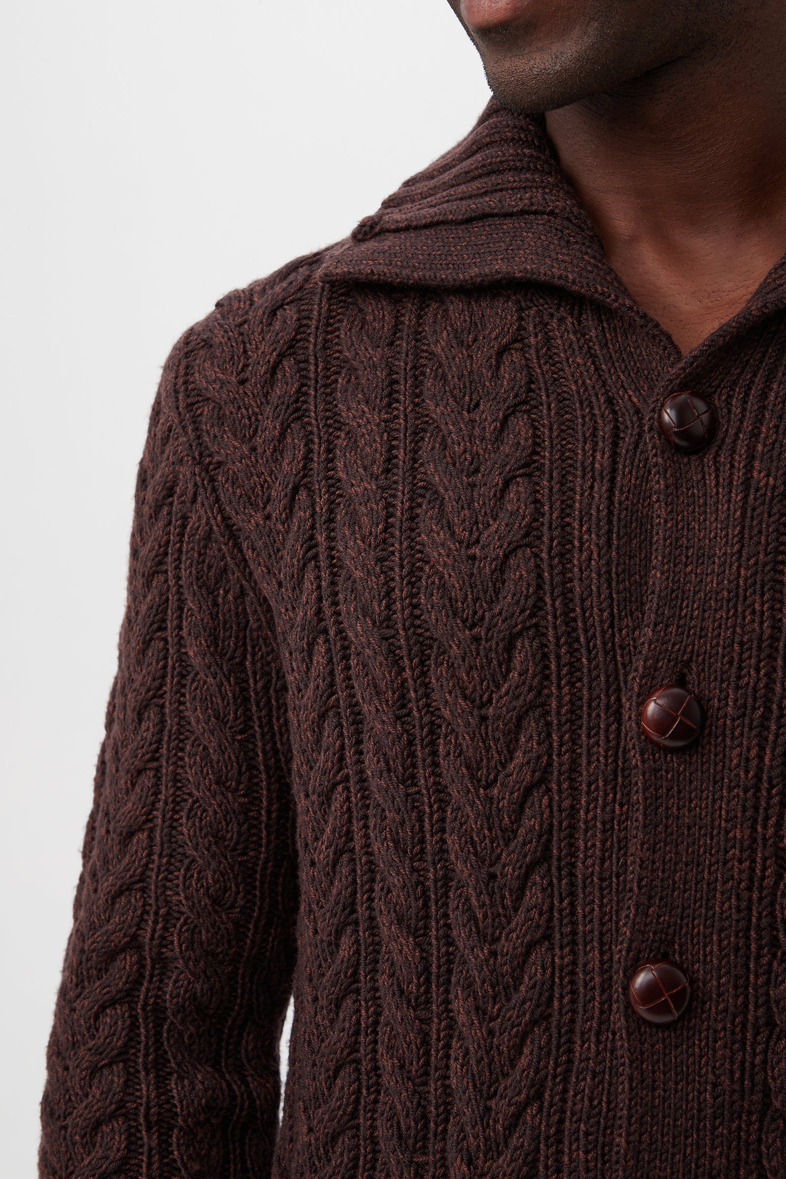 A person wears the Raleigh Sweater Coat – Merino Wool, a brown cable-knit cardigan with large buttons and a wide collar. Their face is partially visible against a plain white background.