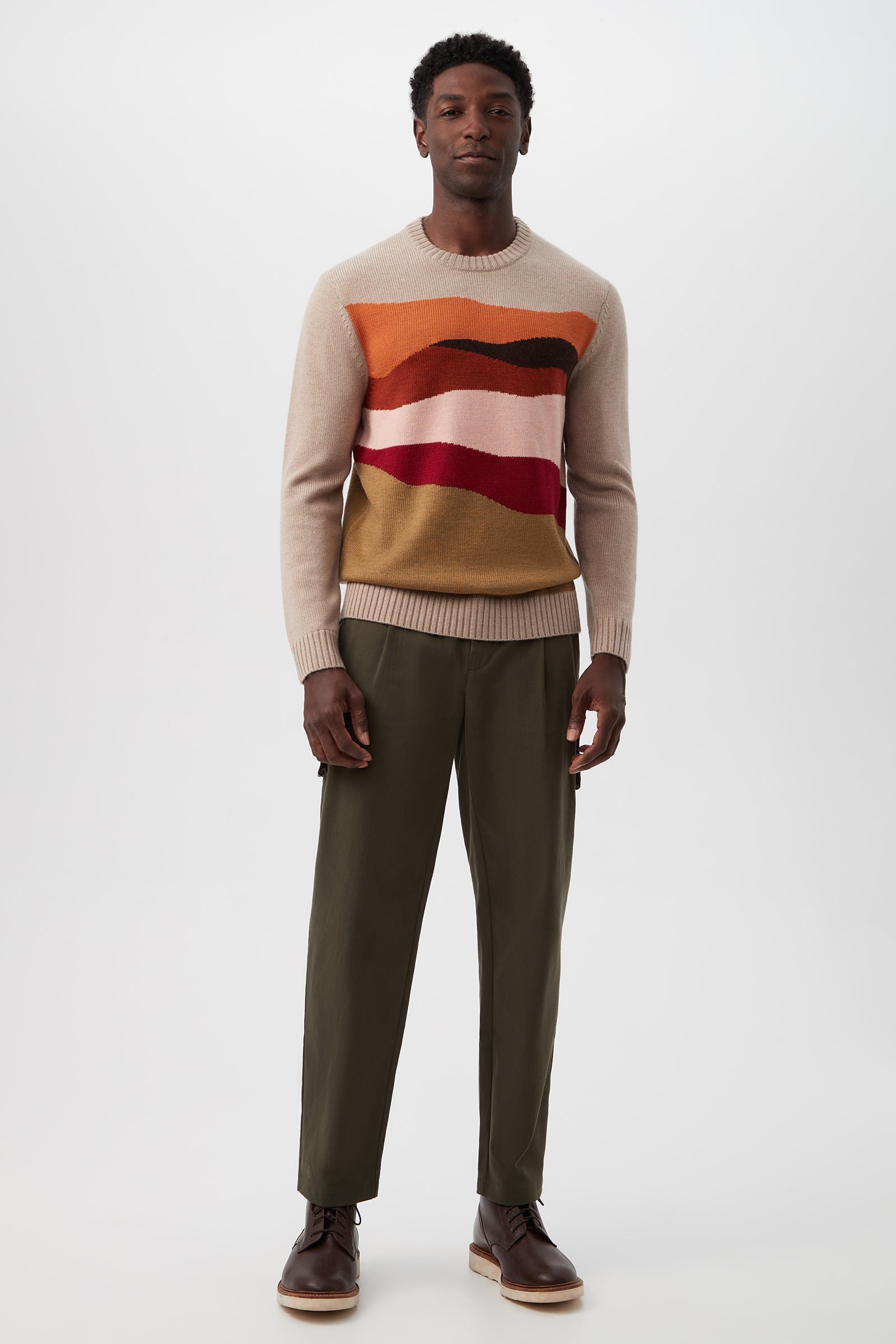 A man stands against a plain background wearing the Murphy Long-Sleeve Crewneck - Merino Wool, featuring wavy red, orange, and burgundy stripes, paired with olive green pants and brown boots. He looks relaxed with his arms at his sides.