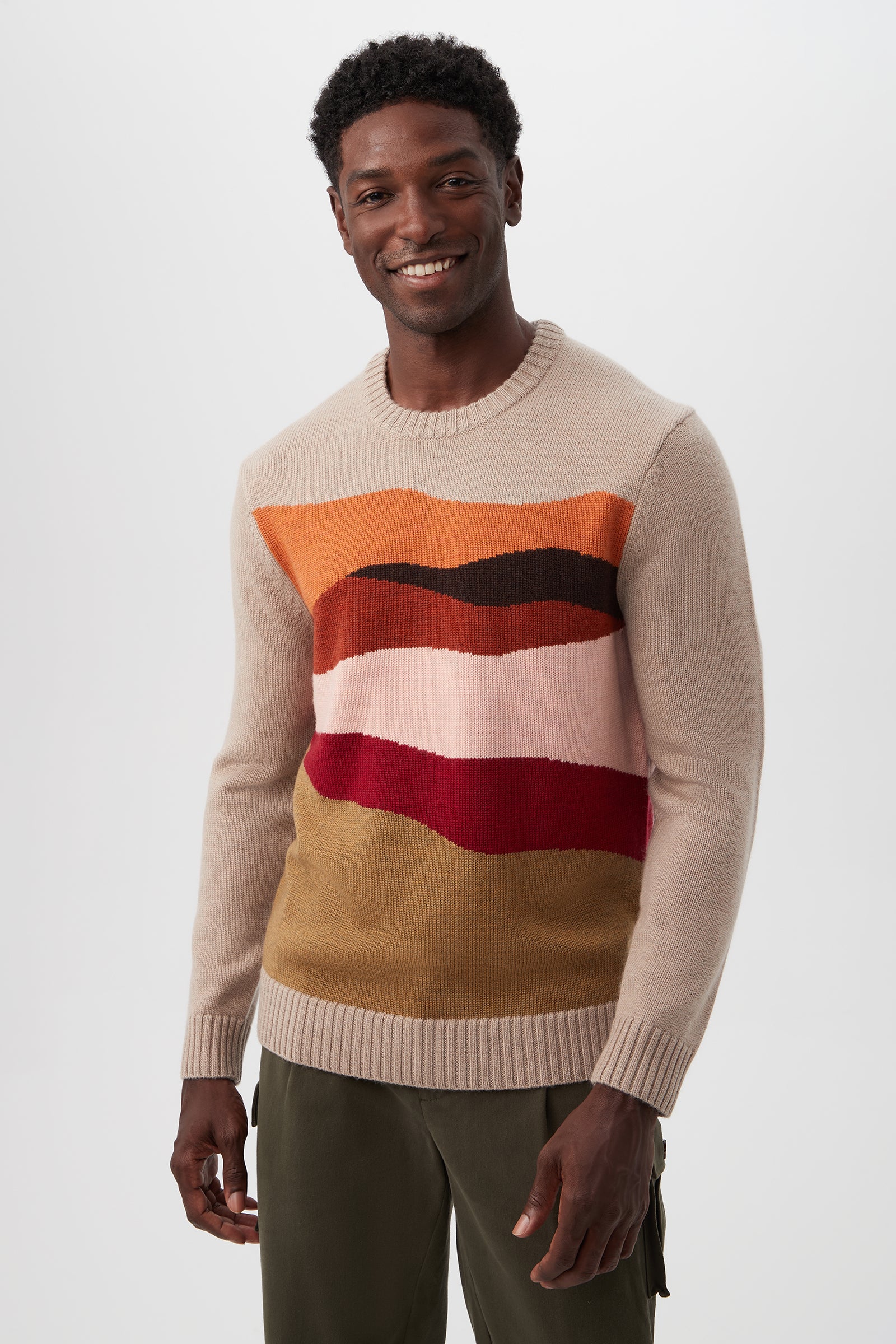 A man with short, curly hair smiles while wearing the Murphy Long-Sleeve Crewneck - Merino Wool in Camel Multi, featuring colorful wavy stripes on the chest and paired with olive green pants against a plain white background.