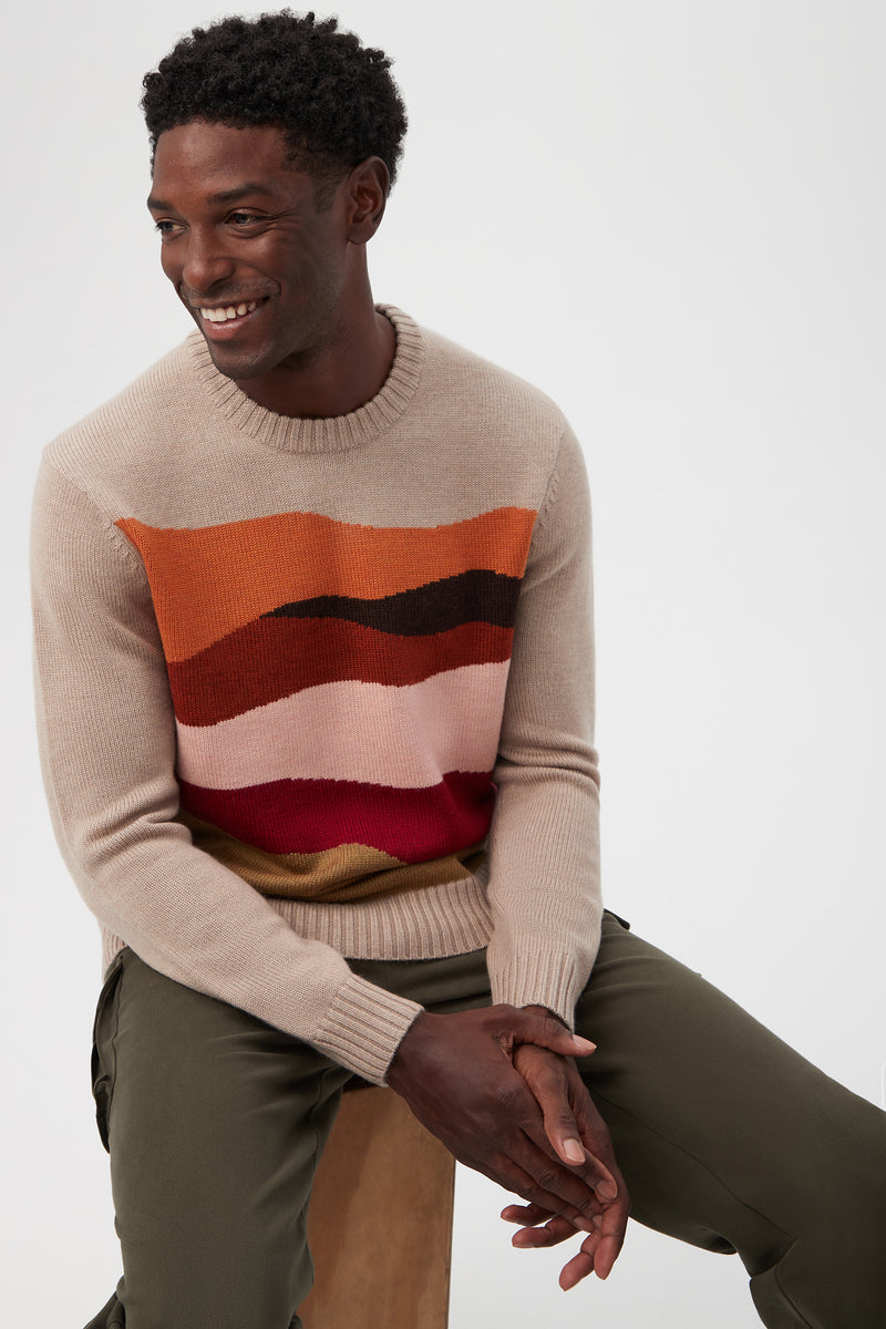 A smiling man wears the Murphy Long-Sleeve Crewneck in beige merino wool with red, orange, pink, and brown stripes, paired with olive green pants. He sits on a wooden stool against a plain light background.