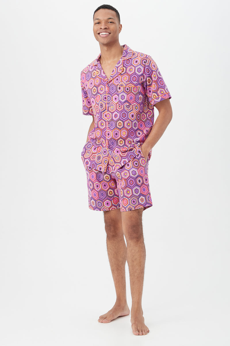 A smiling man stands barefoot against a white background, wearing the Honeycomb Dots Mens Short Sleeve Boxer Short Jersey PJ Set with a colorful geometric pattern. His hands rest in his pockets, highlighting the short sleeve button-up pajama top.