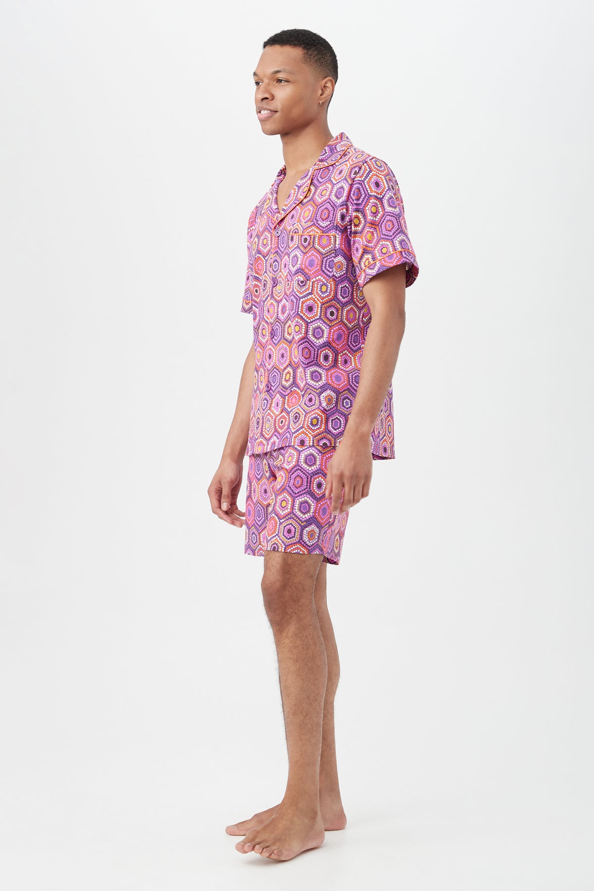 A person stands barefoot on a light background, wearing the Honeycomb Dots Mens Short Sleeve Boxer Short Jersey PJ Set, which features a matching button-up shirt and shorts with a colorful geometric pattern in pink, purple, and orange.