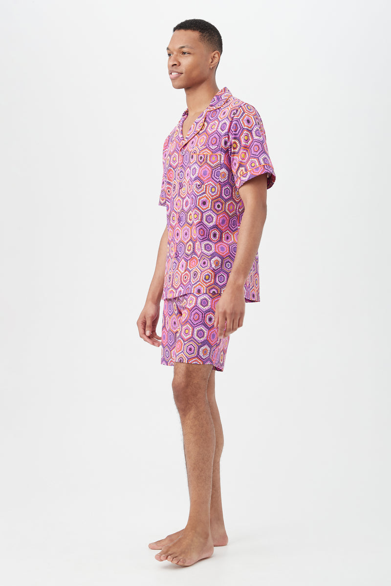 A person stands barefoot on a light background, wearing the Honeycomb Dots Mens Short Sleeve Boxer Short Jersey PJ Set, which features a matching button-up shirt and shorts with a colorful geometric pattern in pink, purple, and orange.