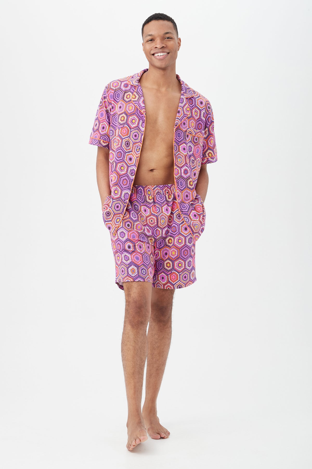 A person stands barefoot against a white background, smiling and wearing the Honeycomb Dots Mens Short Sleeve Boxer Short Jersey PJ Set with a pink and purple geometric pattern, short sleeve button-up top and matching shorts, hands in pockets.
