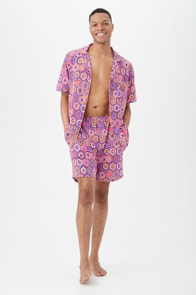 A person stands barefoot against a white background, smiling and wearing the Honeycomb Dots Mens Short Sleeve Boxer Short Jersey PJ Set with a pink and purple geometric pattern, short sleeve button-up top and matching shorts, hands in pockets.