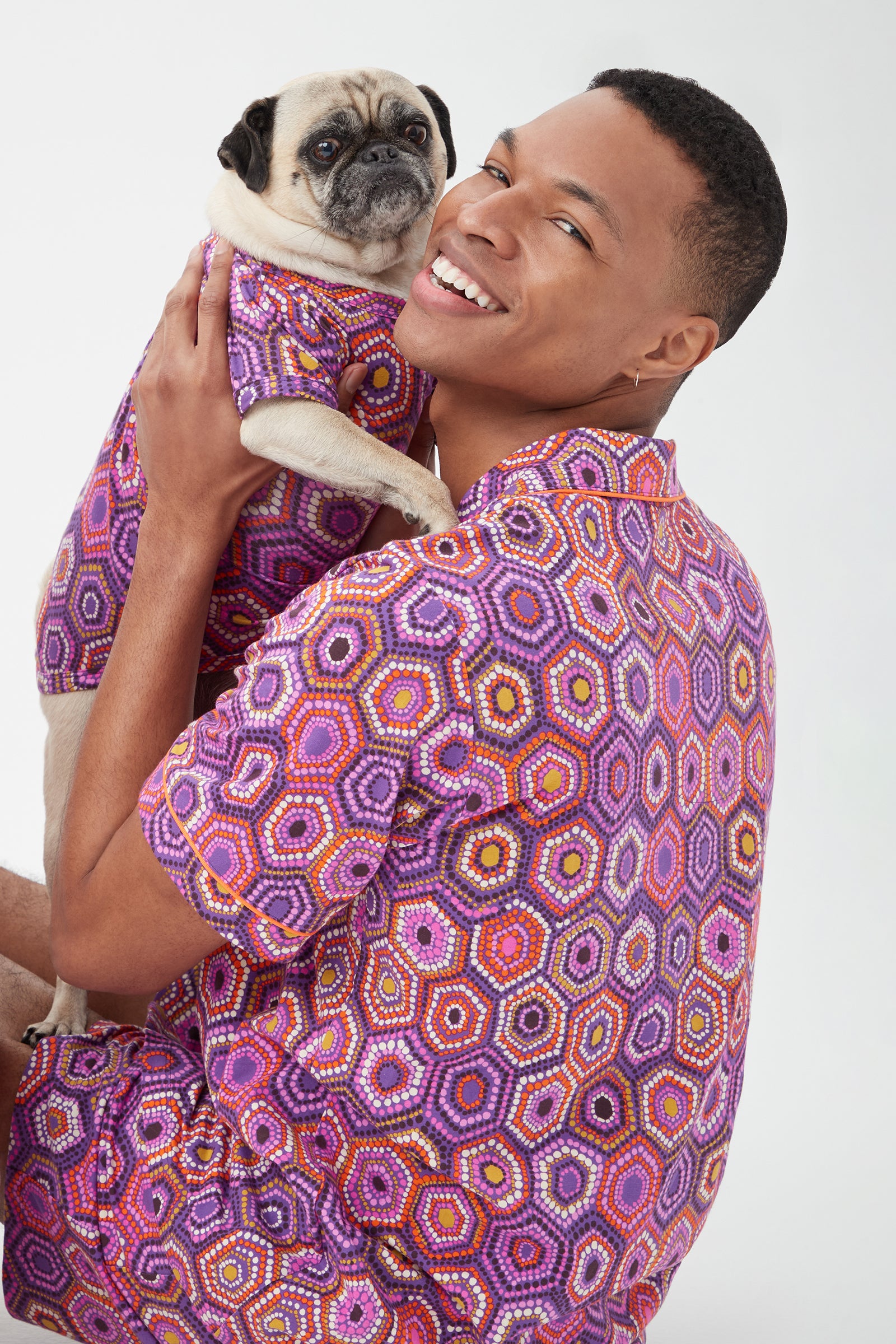 A smiling person holds a pug, both wearing matching Honeycomb Dots Short Sleeve Jersey Dog Pajama Shirts with vibrant geometric patterns in purple, pink, and orange against a plain white background.