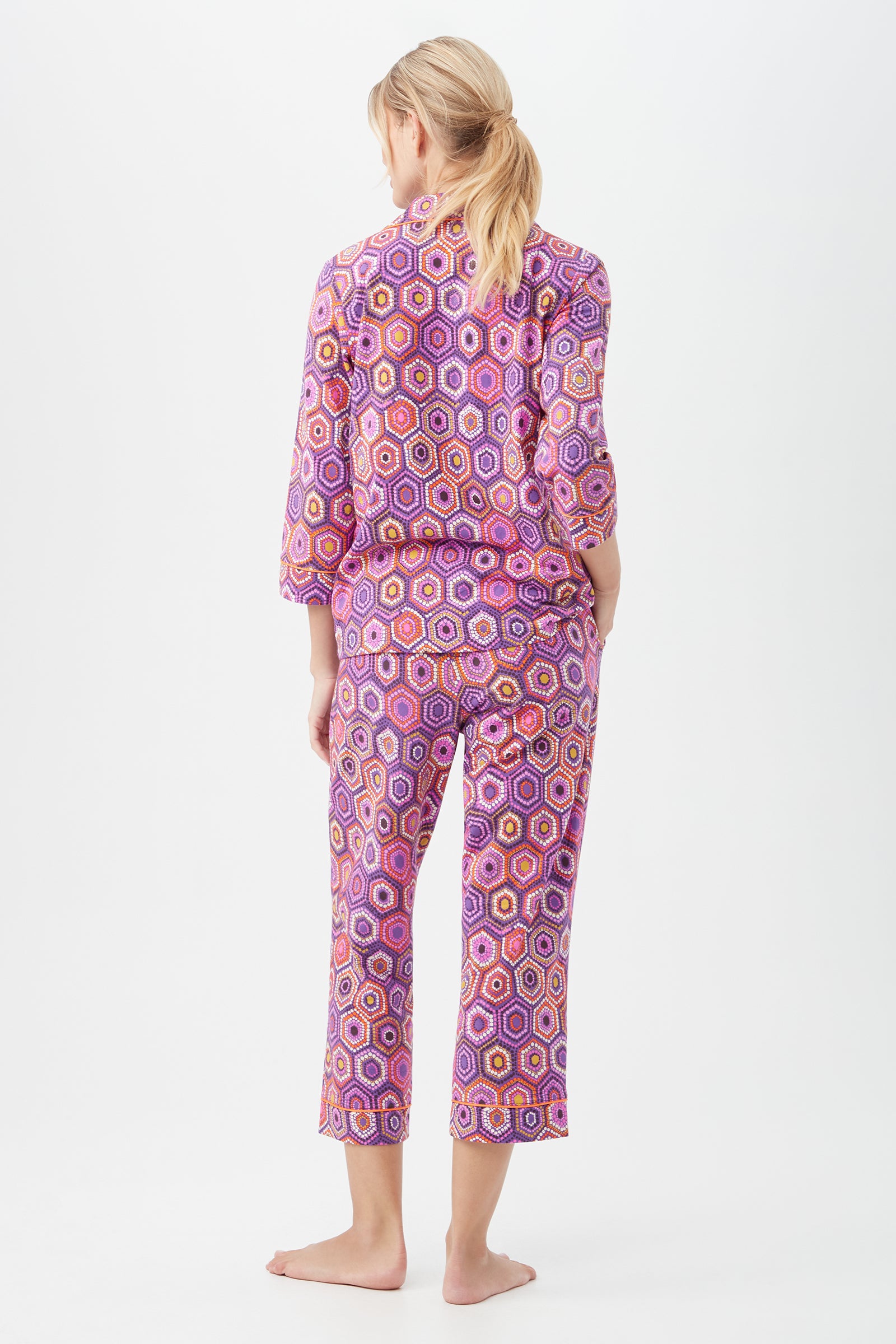 A barefoot woman with blonde hair in a ponytail stands facing away, wearing the Honeycomb Dots Womens 3/4-Sleeve Cropped Pant Jersey PJ Set in purple with a multicolored hexagonal pattern. The background is plain white.