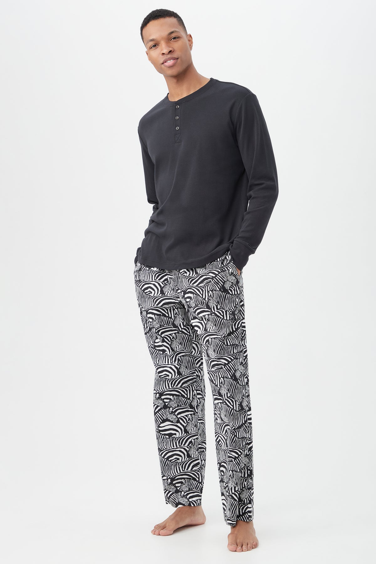 A man stands barefoot against a white background, wearing the Zebra Safari Mens Long Sleeve Henley Long Pant Poplin PJ Setâ€â€featuring a black henley shirt and black-and-white patterned pajama pants, hands in pockets and smiling slightly.