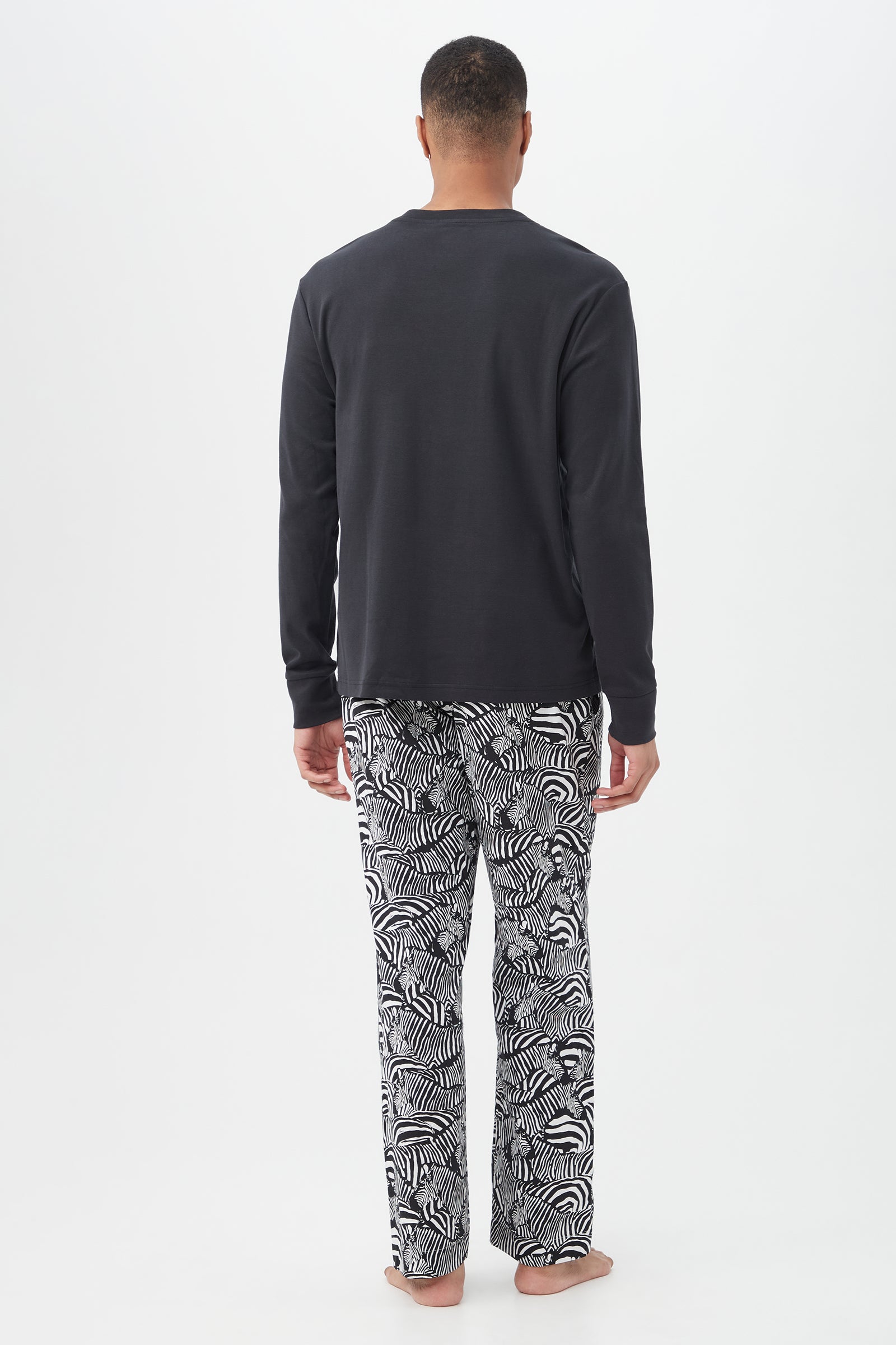 A man stands barefoot, facing away, wearing the Zebra Safari Mens Long Sleeve Henley Long Pant Poplin PJ Set—featuring a black long-sleeve shirt and loose white pajama pants with a black zebra pattern—against a plain white background.
