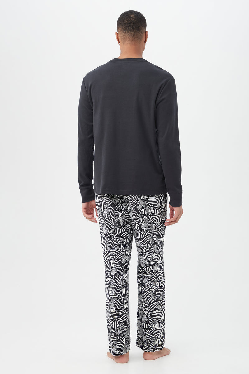 A man stands barefoot, facing away, wearing the Zebra Safari Mens Long Sleeve Henley Long Pant Poplin PJ Setâ€â€featuring a black long-sleeve shirt and loose white pajama pants with a black zebra patternâ€â€against a plain white background.