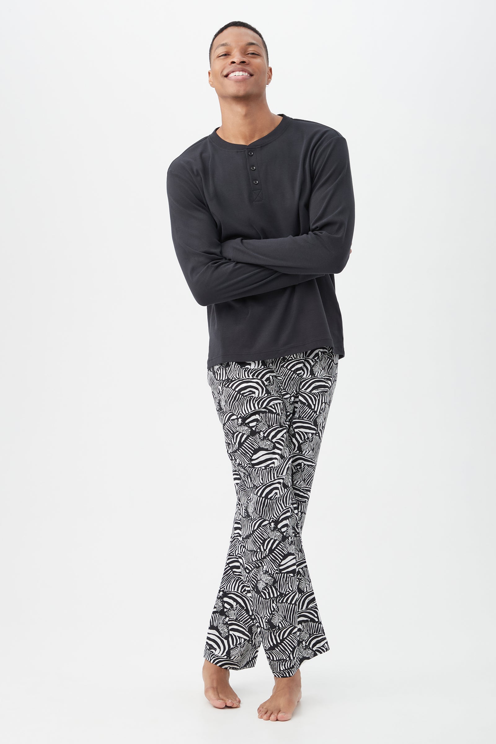A smiling person stands barefoot with arms crossed, wearing a long-sleeved black shirt and the Zebra Safari Mens Long Sleeve Henley Long Pant Poplin PJ Set—a black-and-white patterned mens sleepwear set—against a plain white background.
