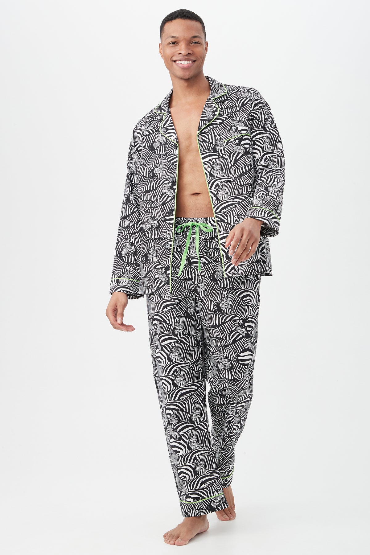 A smiling person walks barefoot in the Zebra Safari Mens Long Sleeve Long Pant Poplin PJ Set—featuring a black-and-white pattern with green trim and drawstring. The lightweight sleepwear top is open, revealing their chest, against a plain white background.